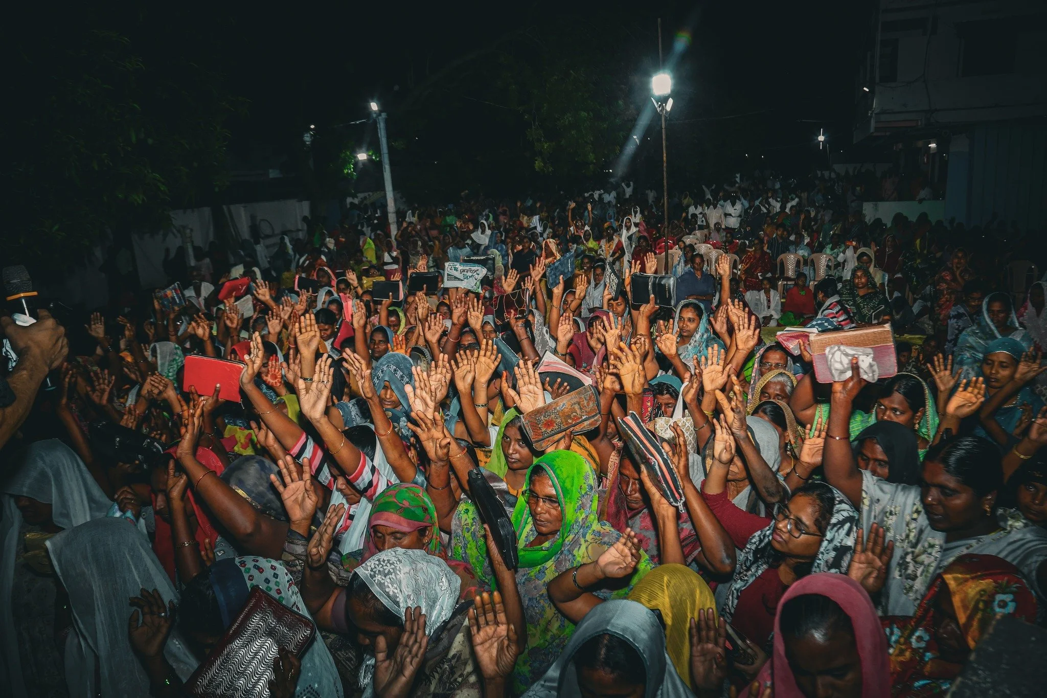 Large crowd of women raising their hands outdoors at night, some holding phones and signs, under bright lights.