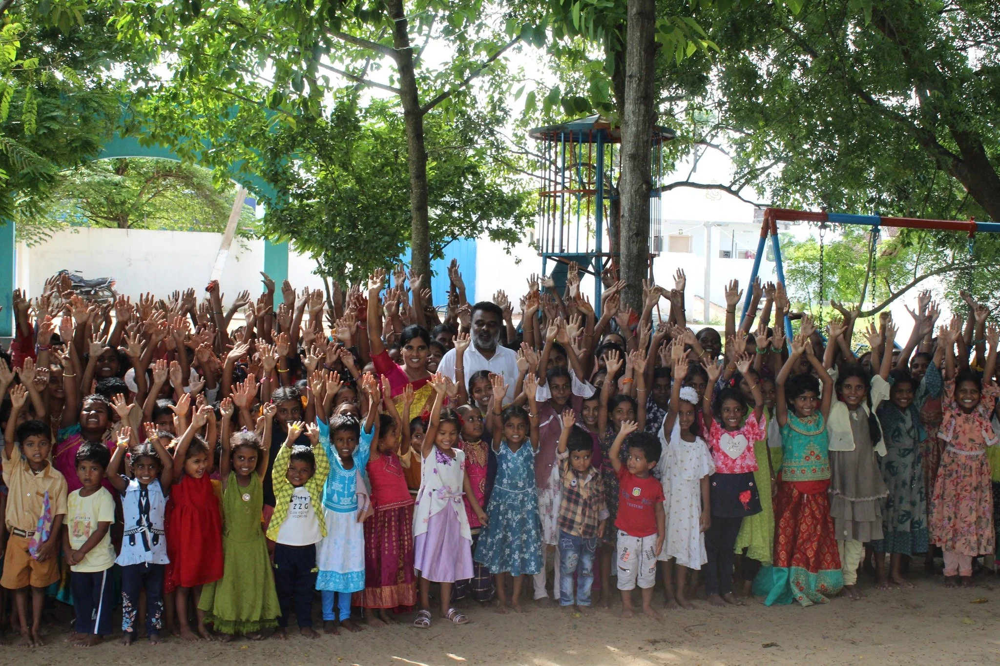 A large group of children and two adults standing outdoors under trees, raising their hands in the air, with playground equipment in the background.