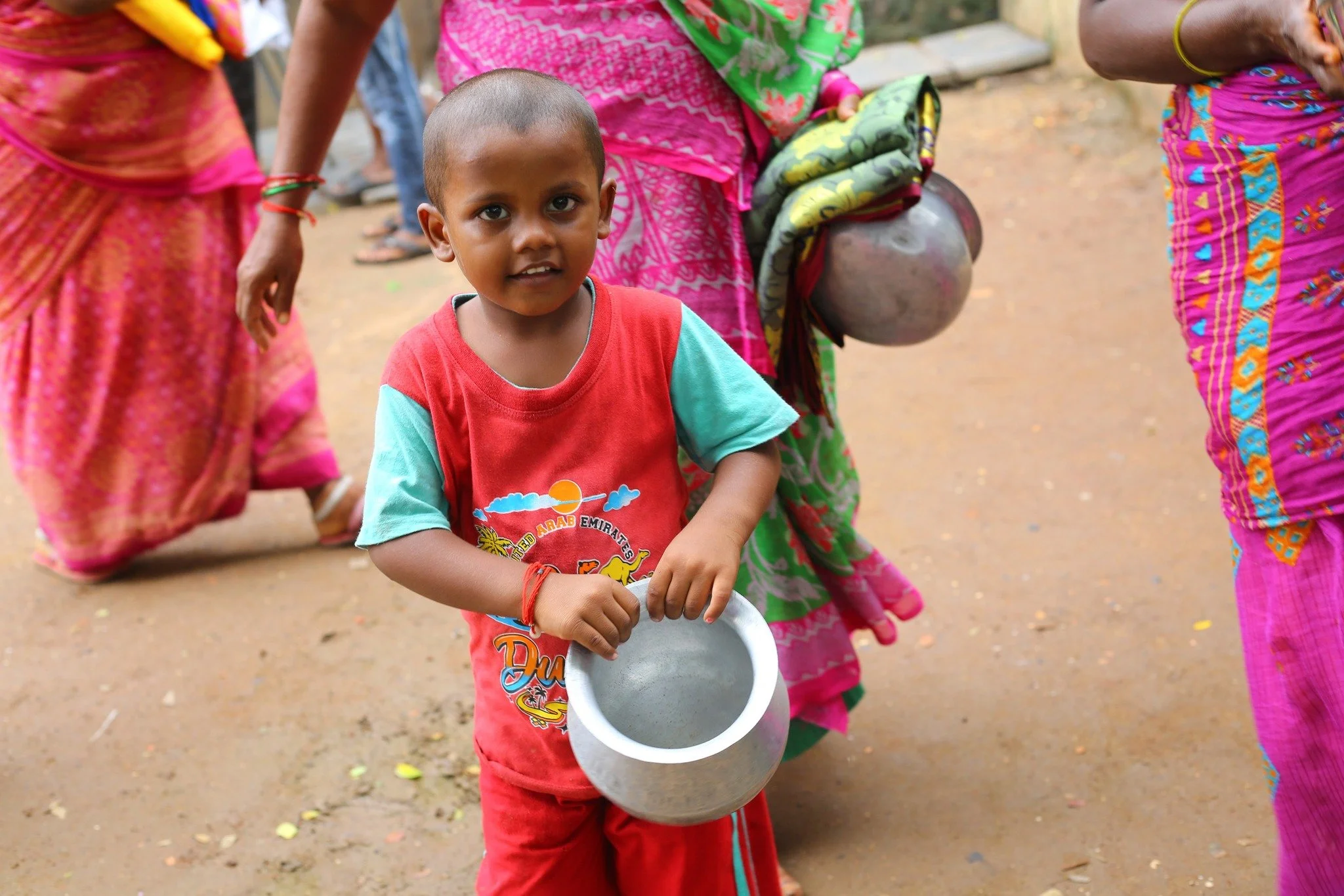 A young boy holding an empty metal bowl, standing on a dirt path surrounded by women dressed in colorful traditional clothing.