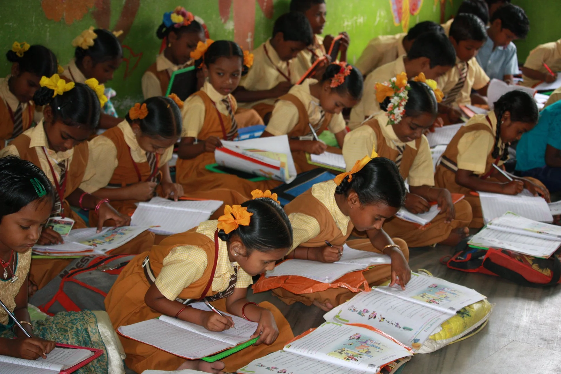 Group of young students sitting on the floor, working in books, in a classroom with green walls decorated with drawings and artwork. The girls are wearing school uniforms with yellow shirts, brown pinafores, and appear to be engaged in reading or writing.