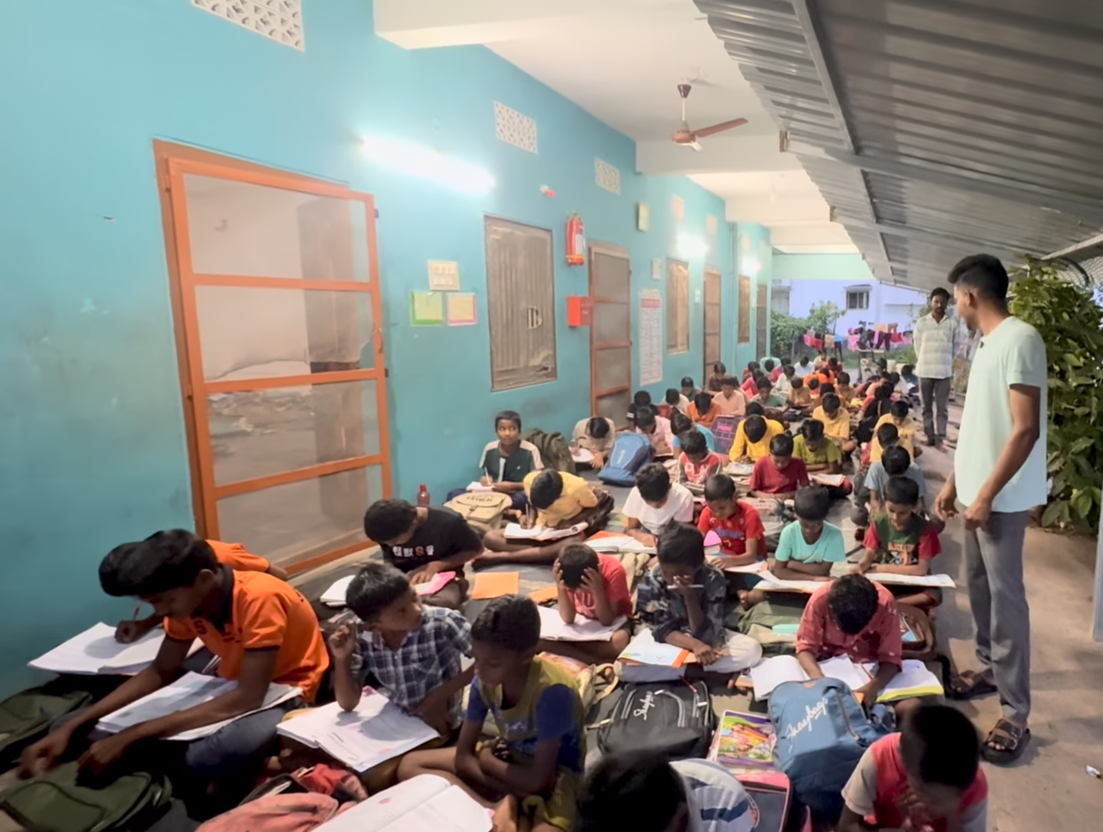 A classroom filled with young students sitting on the floor, studying with notebooks and textbooks, while two teachers supervise.