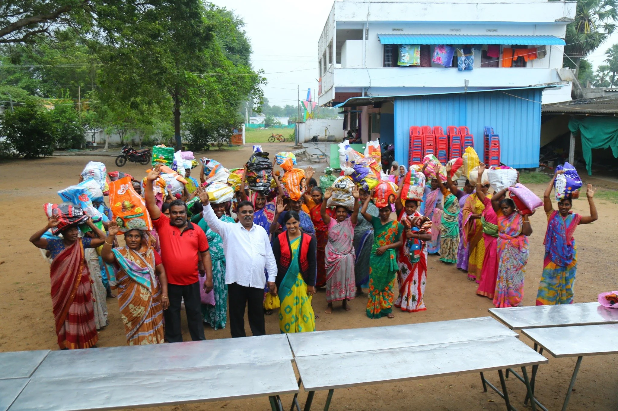 Group of women and men, many wearing colorful saris, standing outdoors with heads and shoulders covered with large plastic bags, some smiling and waving, in a rural setting with a building and trees in the background.