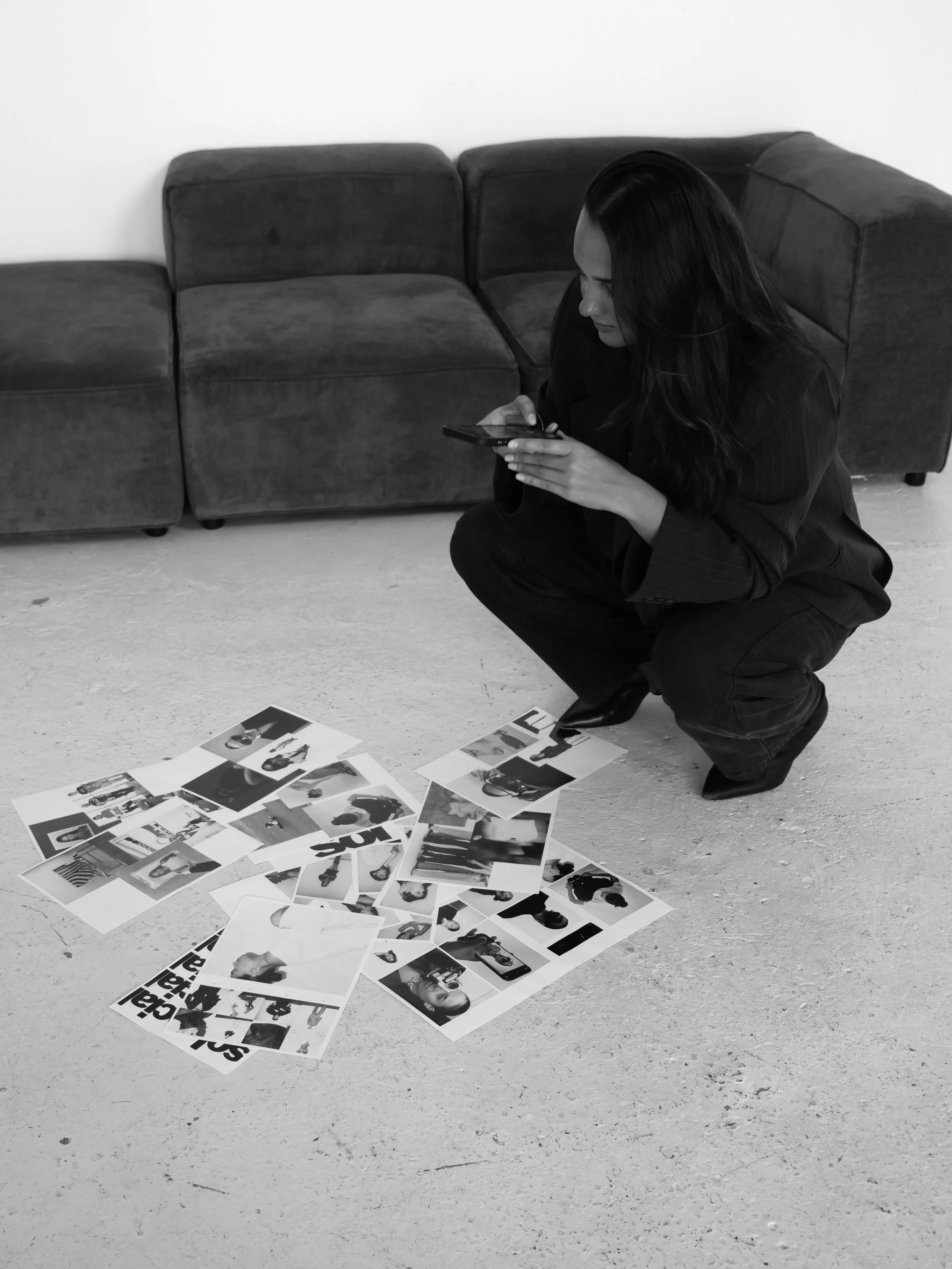 A woman with long dark hair, dressed in black, squatting on the floor and examining photographs spread out in front of her. She is holding a smartphone in her right hand, possibly taking notes or looking at the photos. There is a dark-colored couch behind her against a plain white wall.