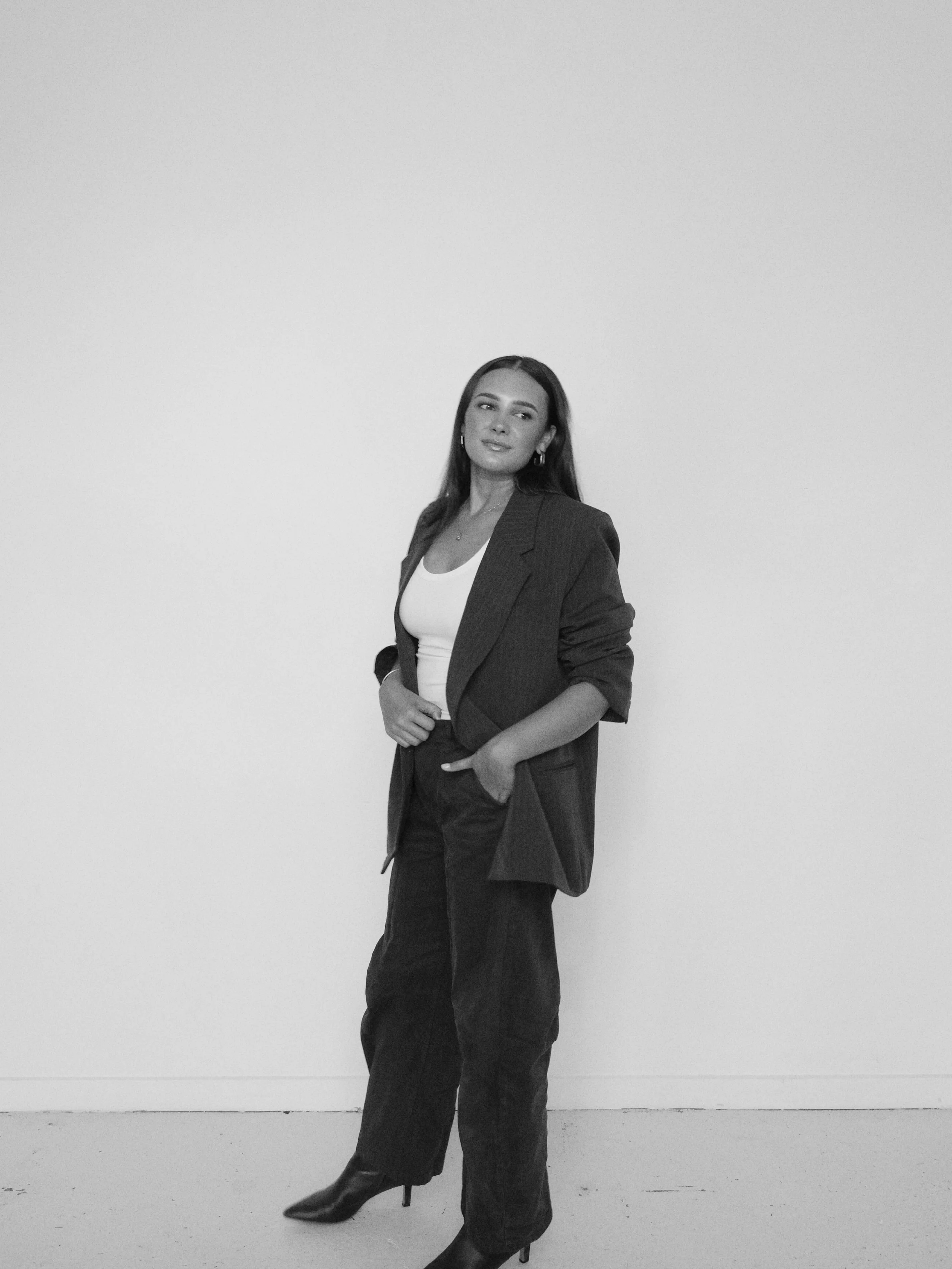 A woman in a blazer, white top, and high heels standing against a plain wall.
