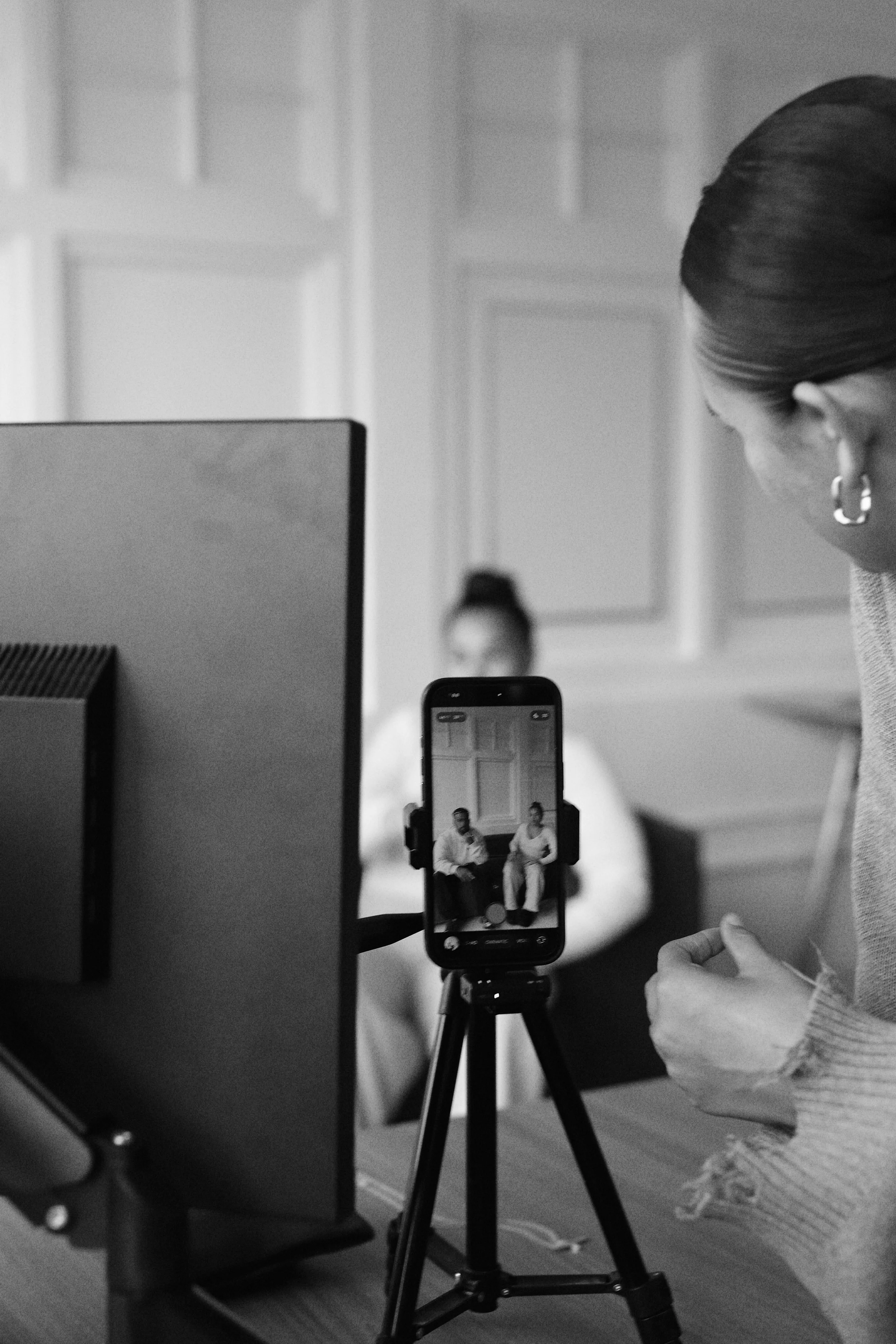 A woman adjusting her clothing while a woman sits in the background during a photo or video recording session in a room with paneled walls. A smartphone on a tripod captures the scene, showing the two women on its screen.