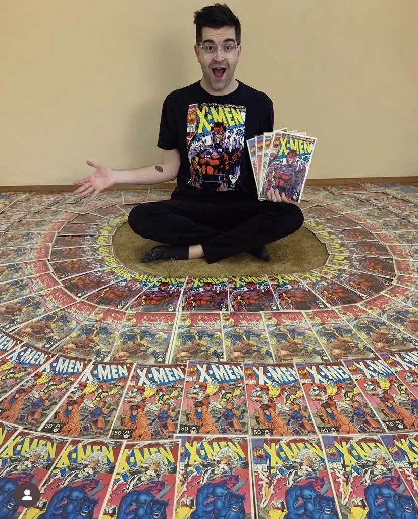 Person sitting on floor surrounded by arranged X-Men comic books, wearing an X-Men T-shirt, holding several issues.