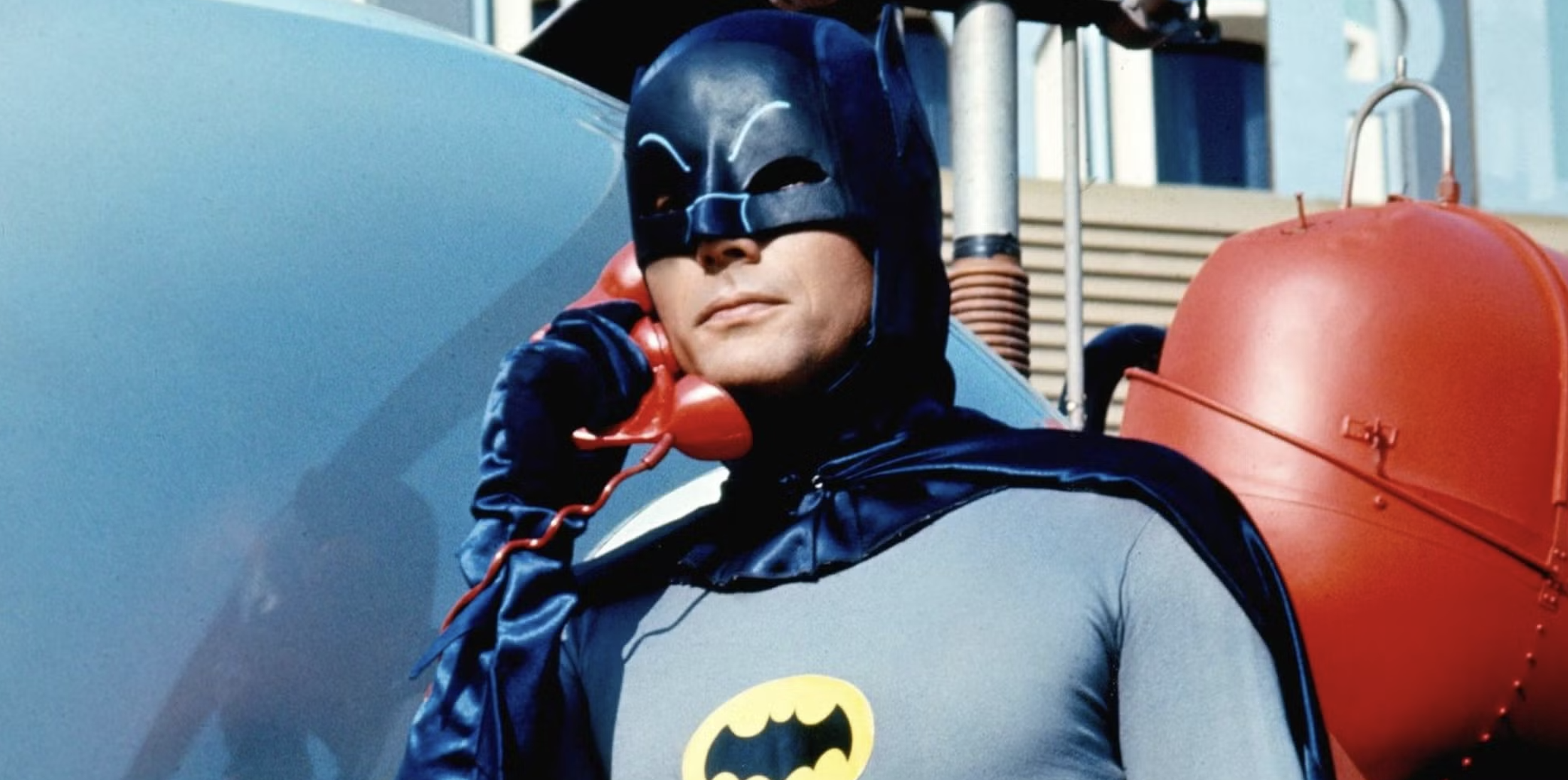 Man in classic superhero costume holding a red phone, wearing a blue mask and cape with a yellow bat symbol on his chest.
