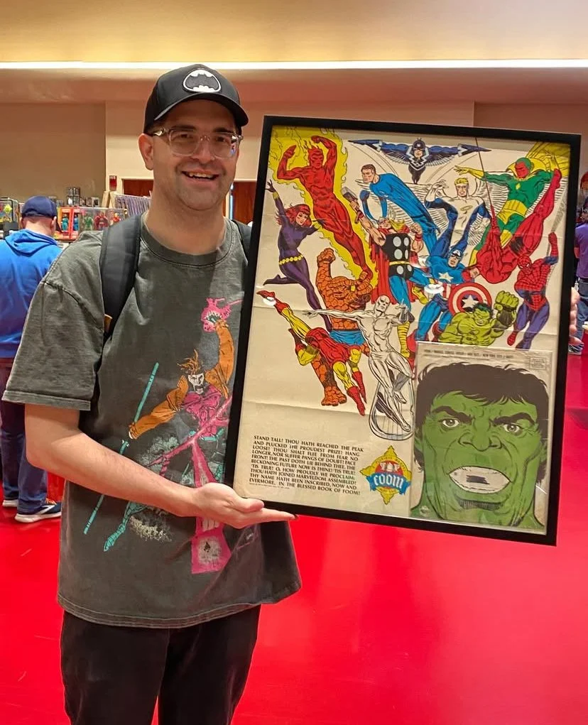 A person wearing a Batman cap and a graphic t-shirt is holding a framed comic art print featuring various superhero characters, including Hulk, Captain America, and Spider-Man, against a red carpet background at a comic convention.