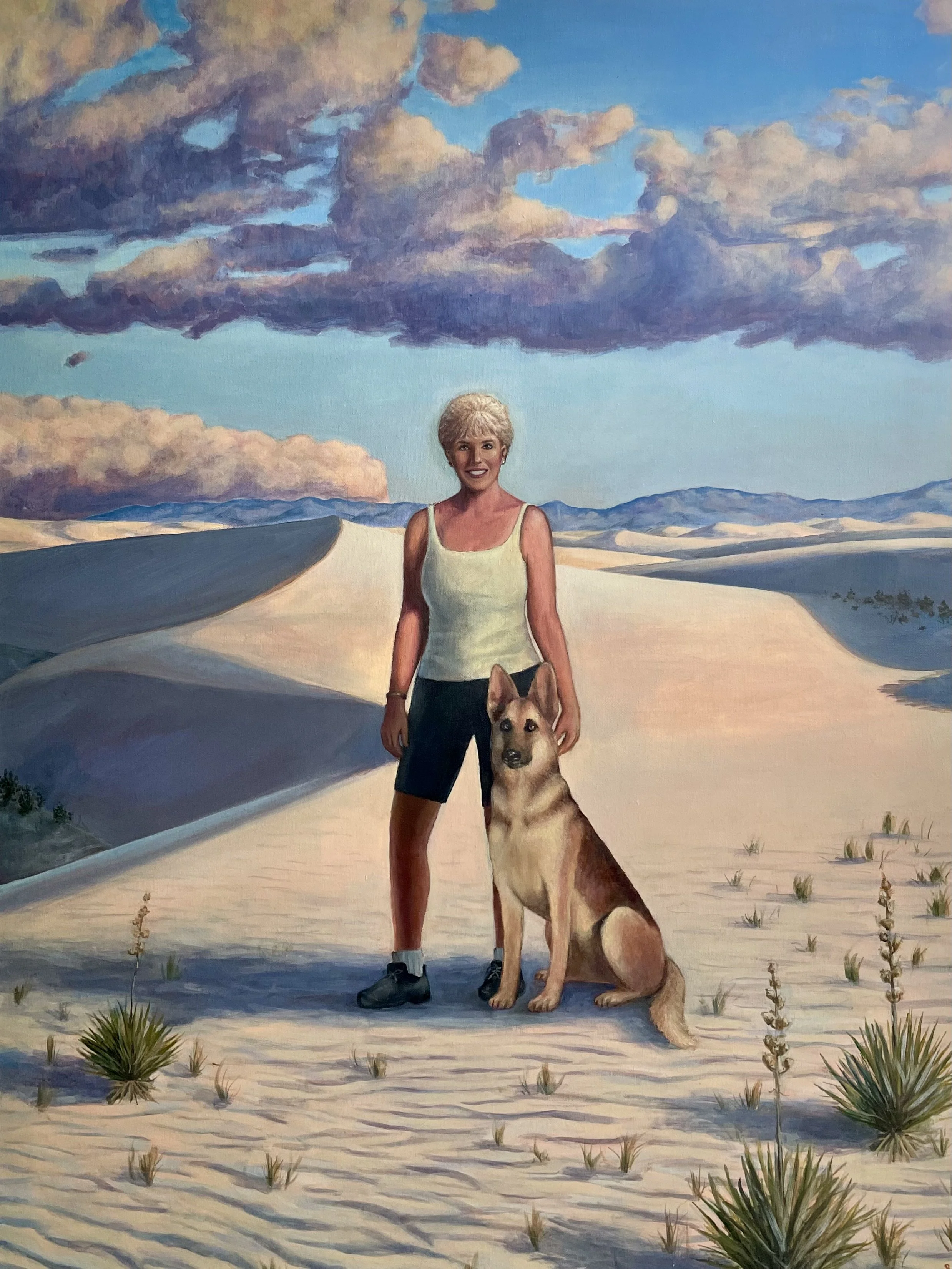 A woman with short blonde hair standing in a desert landscape with a German Shepherd dog.