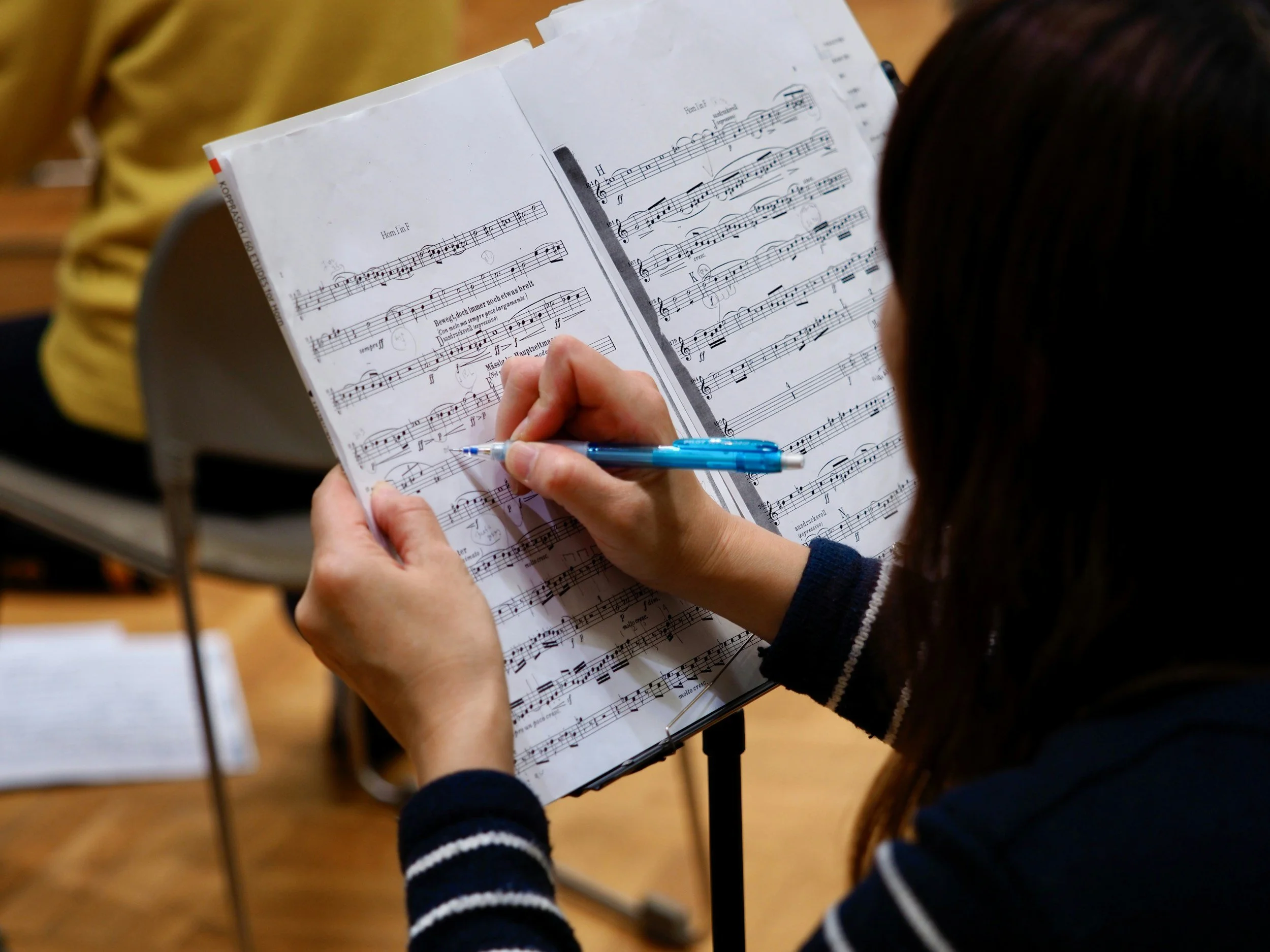 A person reviewing sheet music on a music stand, holding a blue pen, in a room with wooden floors.