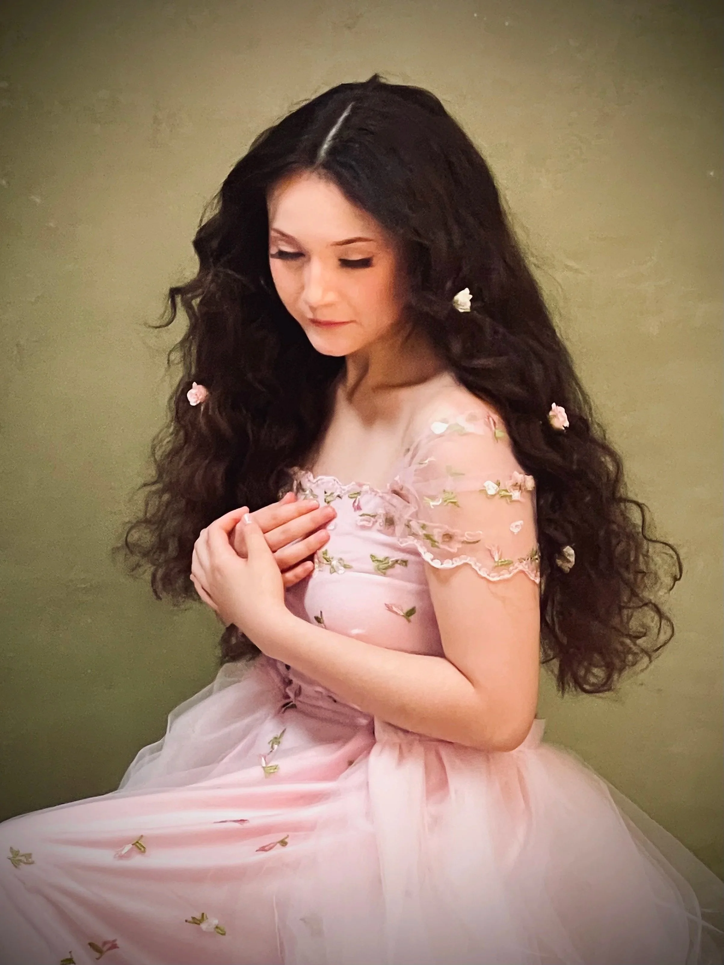 A woman with long, dark, curly hair wearing a pink dress with embroidered floral patterns, sitting against a plain background with flowers in her hair and on her dress, gazing downward with her hands clasped near her chest.