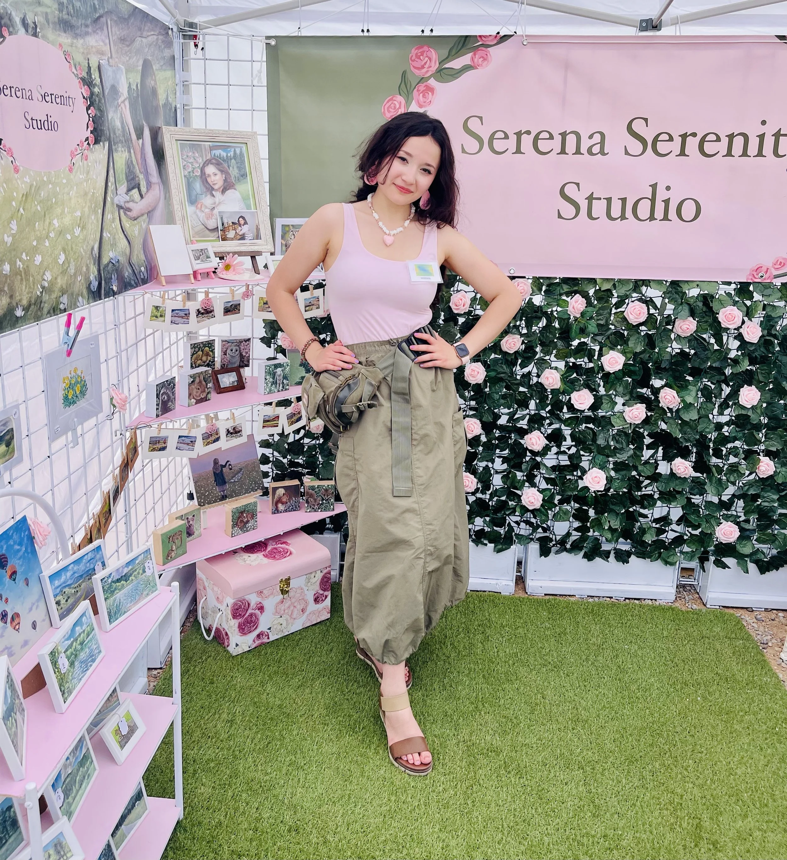 A young woman standing in front of a display at Serena Serenity Studio, surrounded by pink roses and framed photographs, wearing a light pink tank top, olive green baggy pants, and tan sandals.