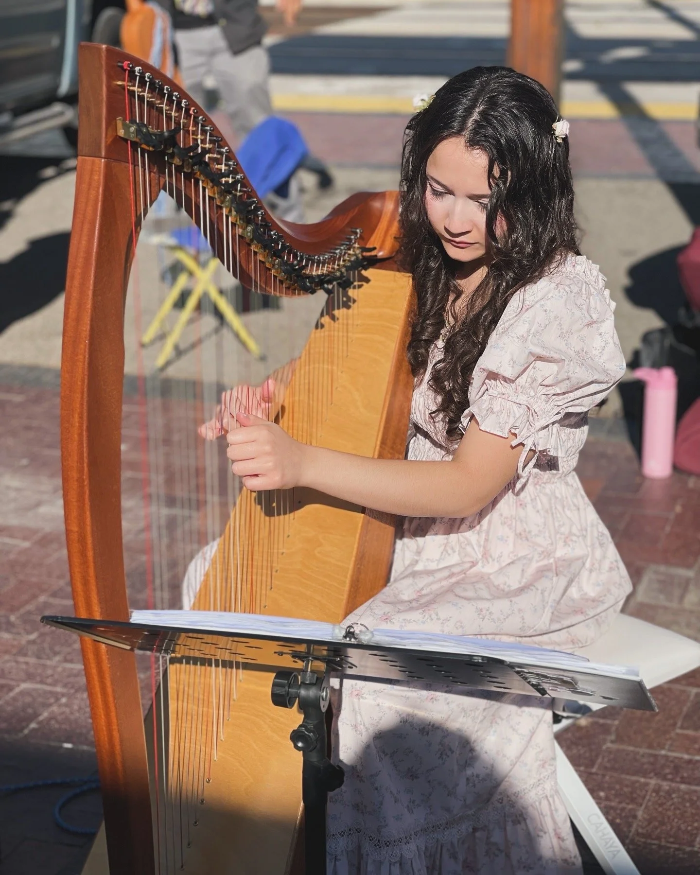 Young girl with long curly dark hair playing a wooden harp, seated outdoors with music sheet on a stand in front of her.