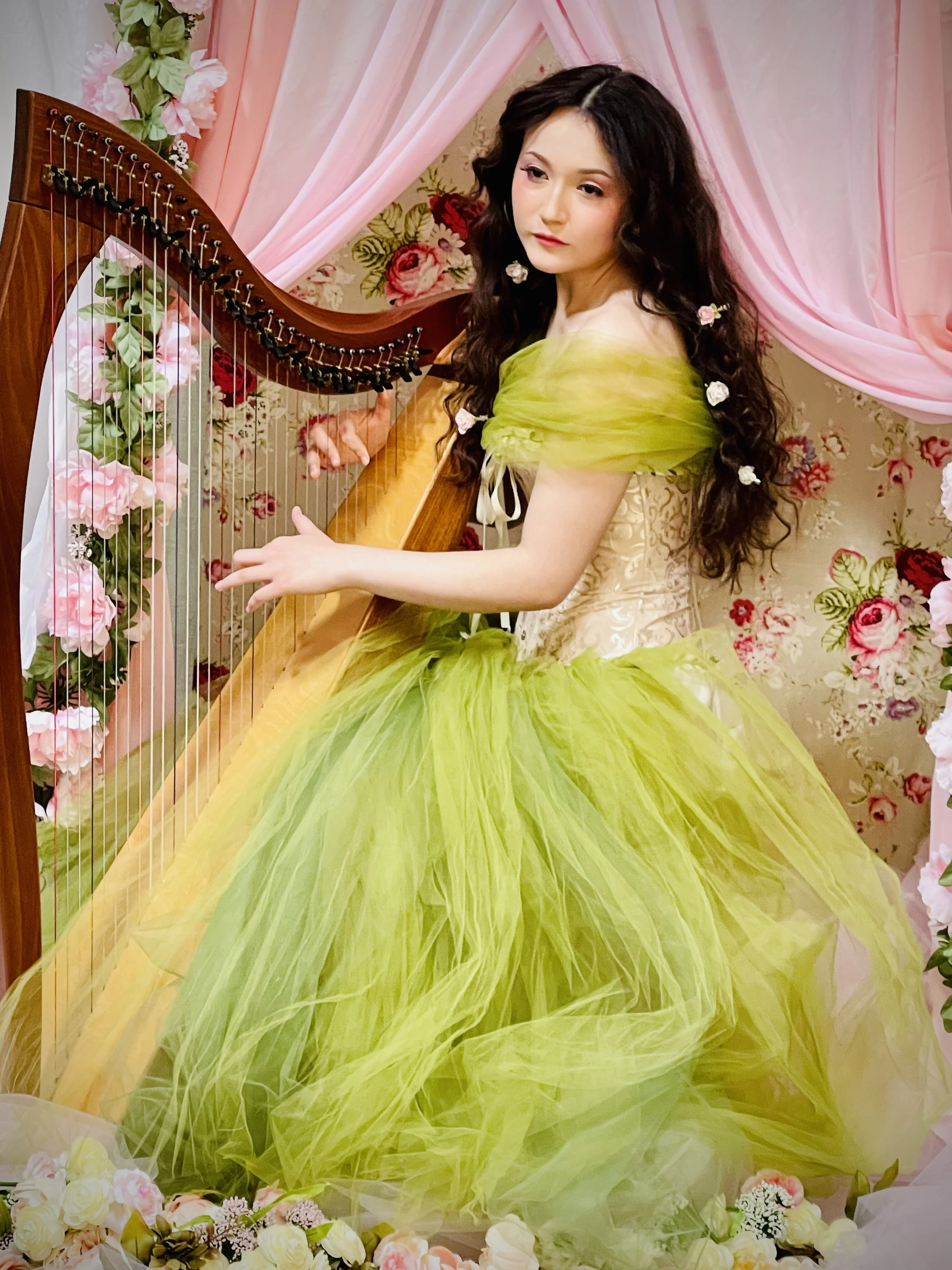 A woman with dark, curly hair wearing a yellow and green gown with tulle skirt, sitting at a harp decorated with roses in a floral and pink draped background.