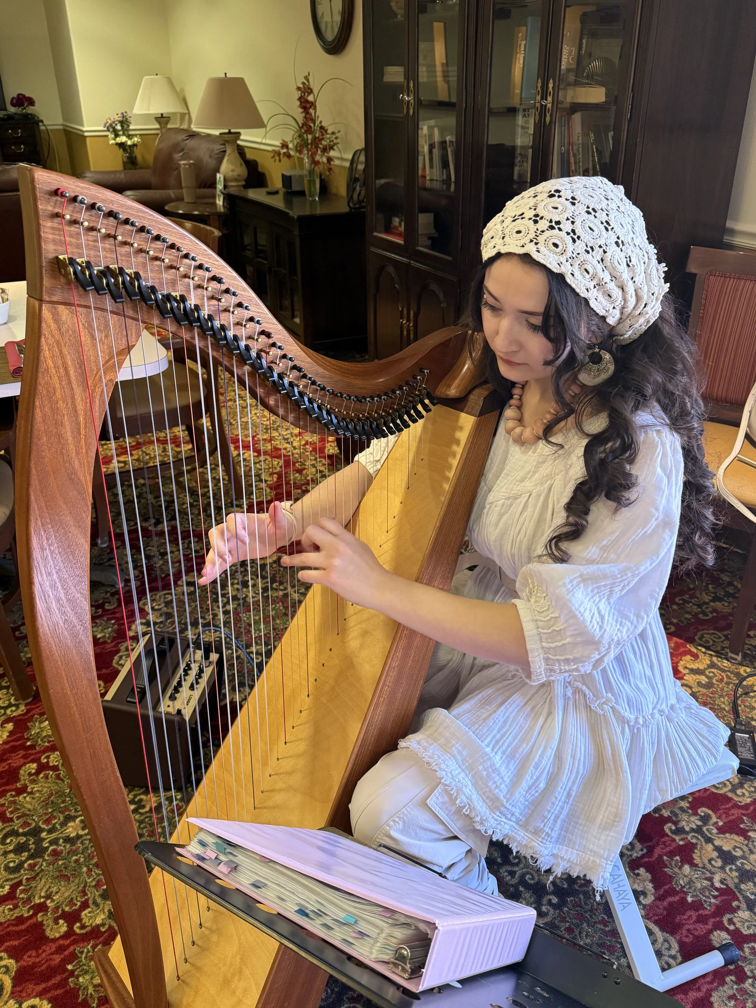 Young woman with long dark curly hair, white crochet hat, and white dress playing a wooden harp in a cozy, decorated room.
