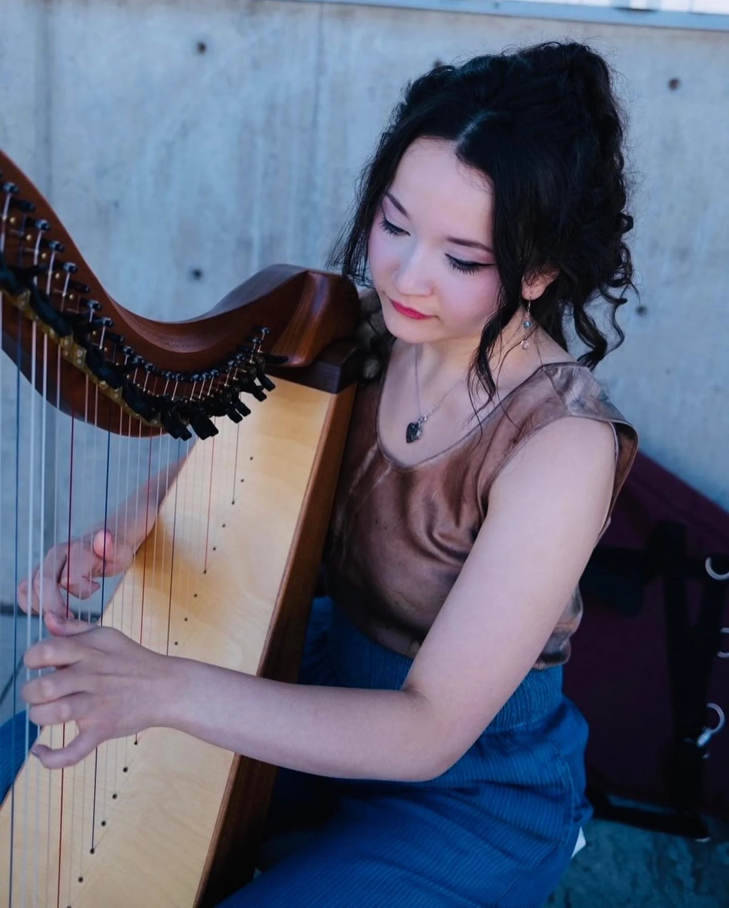 A woman playing a wooden harp. She has curly black hair and is wearing a sleeveless brown top and blue skirt. She is sitting against a concrete wall.