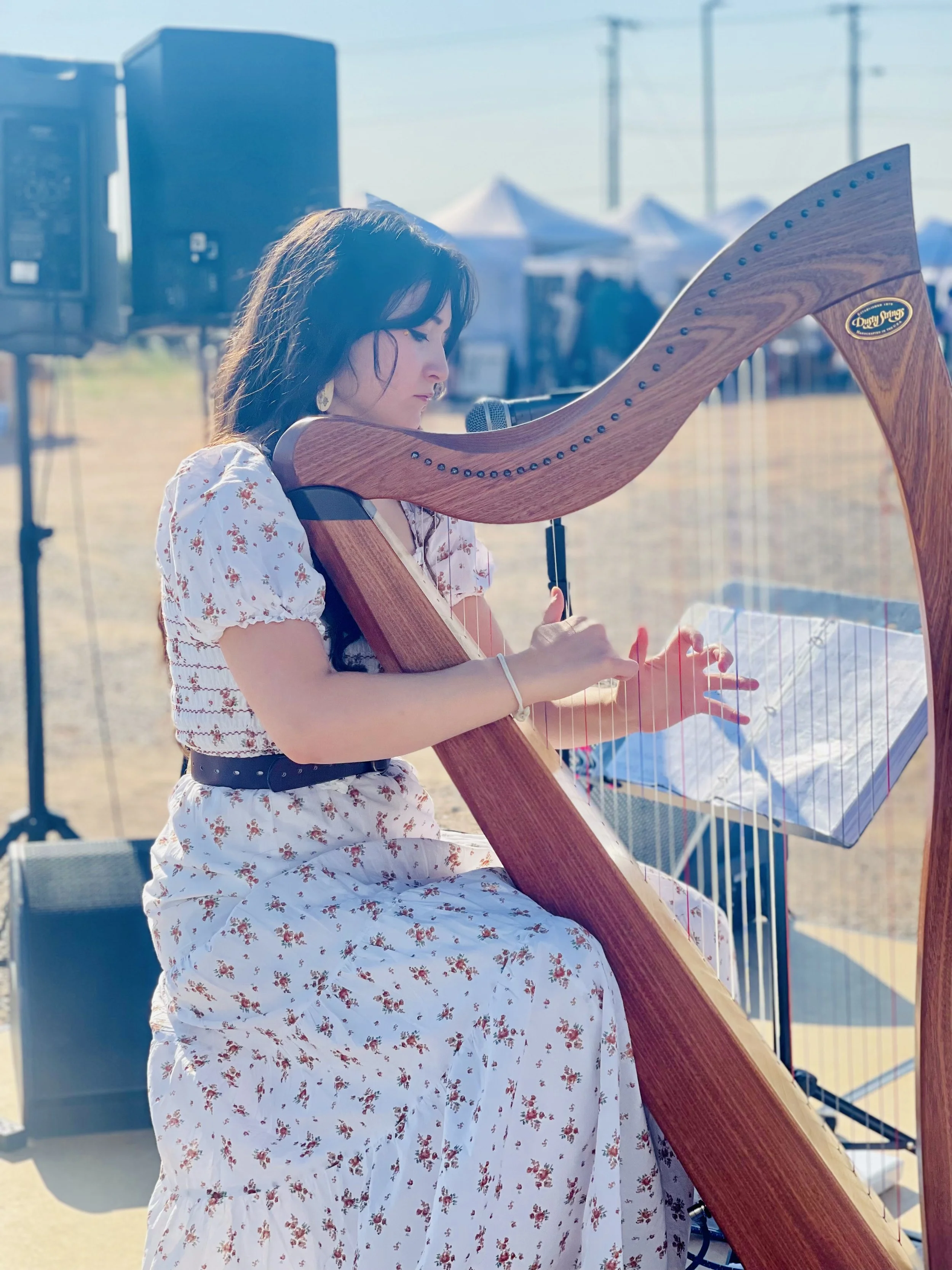 A woman playing a harp at an outdoor event with tents and speakers in the background