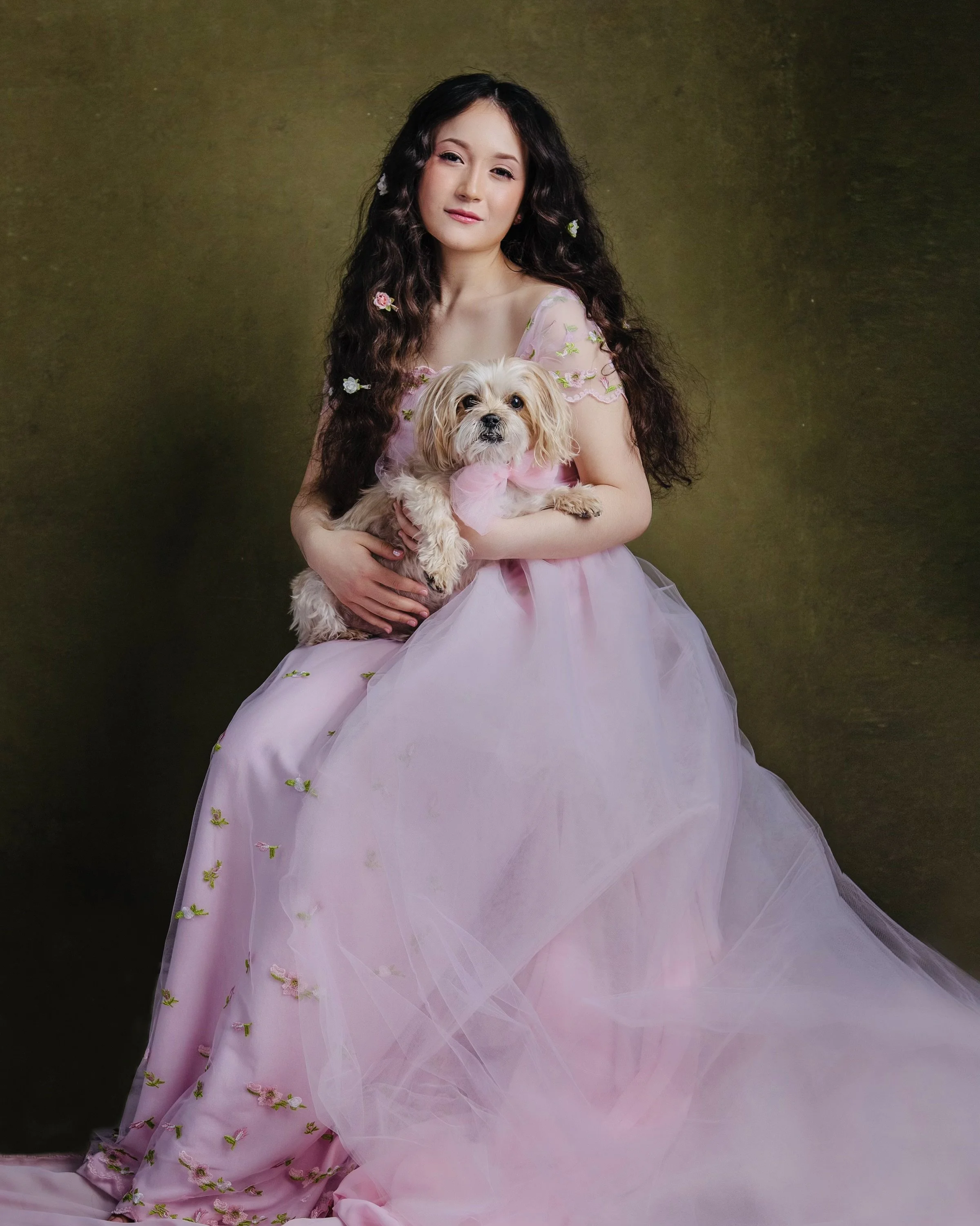 A woman with long dark curly hair wearing a pink dress with embroidery, holding a small dog with light-colored fur and a pink bow, against a dark green background.