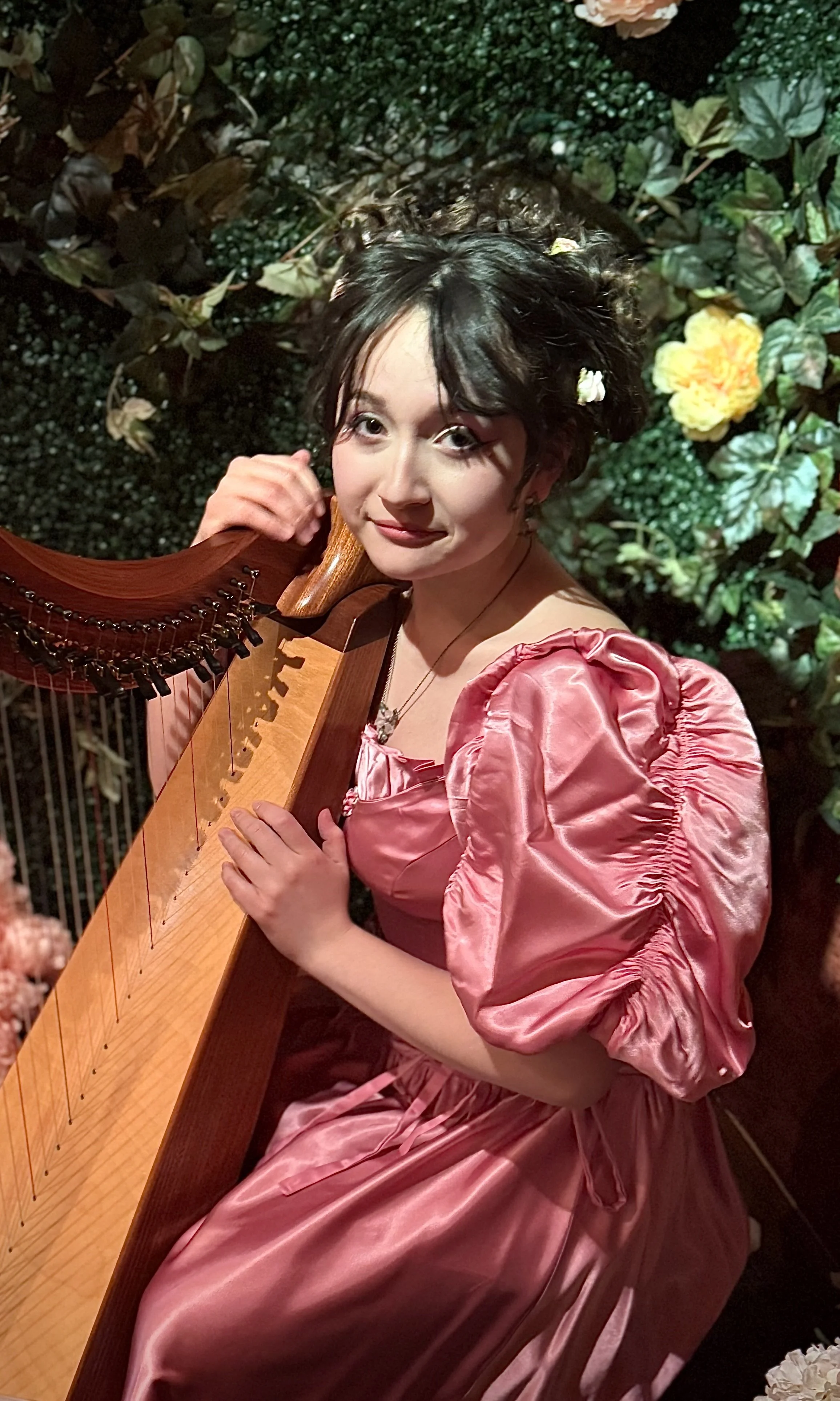 A woman in a pink satin dress with puffed sleeves sitting with a harp in front of a green leafy background with yellow flowers.