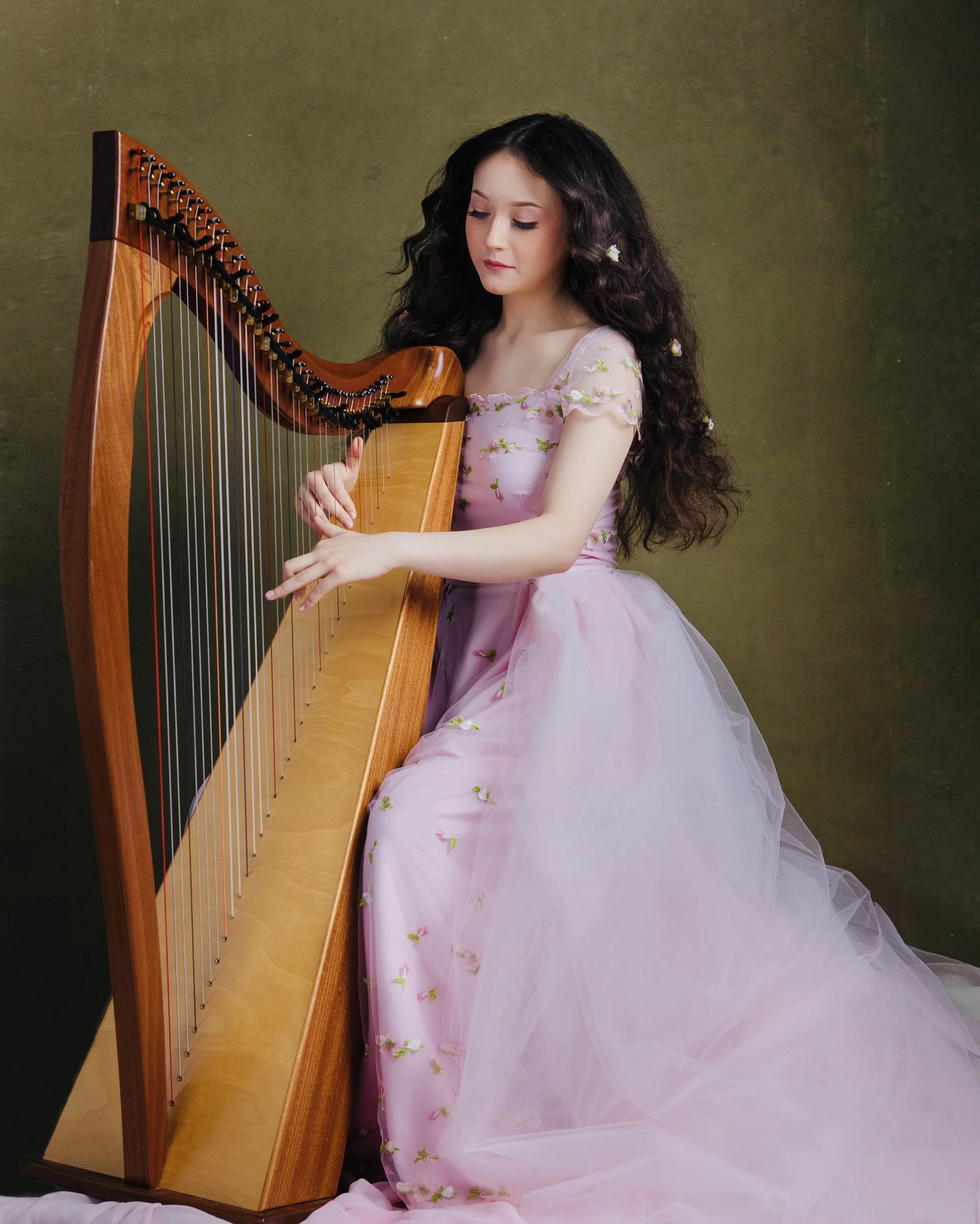 A young woman with long, wavy dark hair dressed in a pink gown with floral embroidery, playing a harp against a plain, dark olive green background.