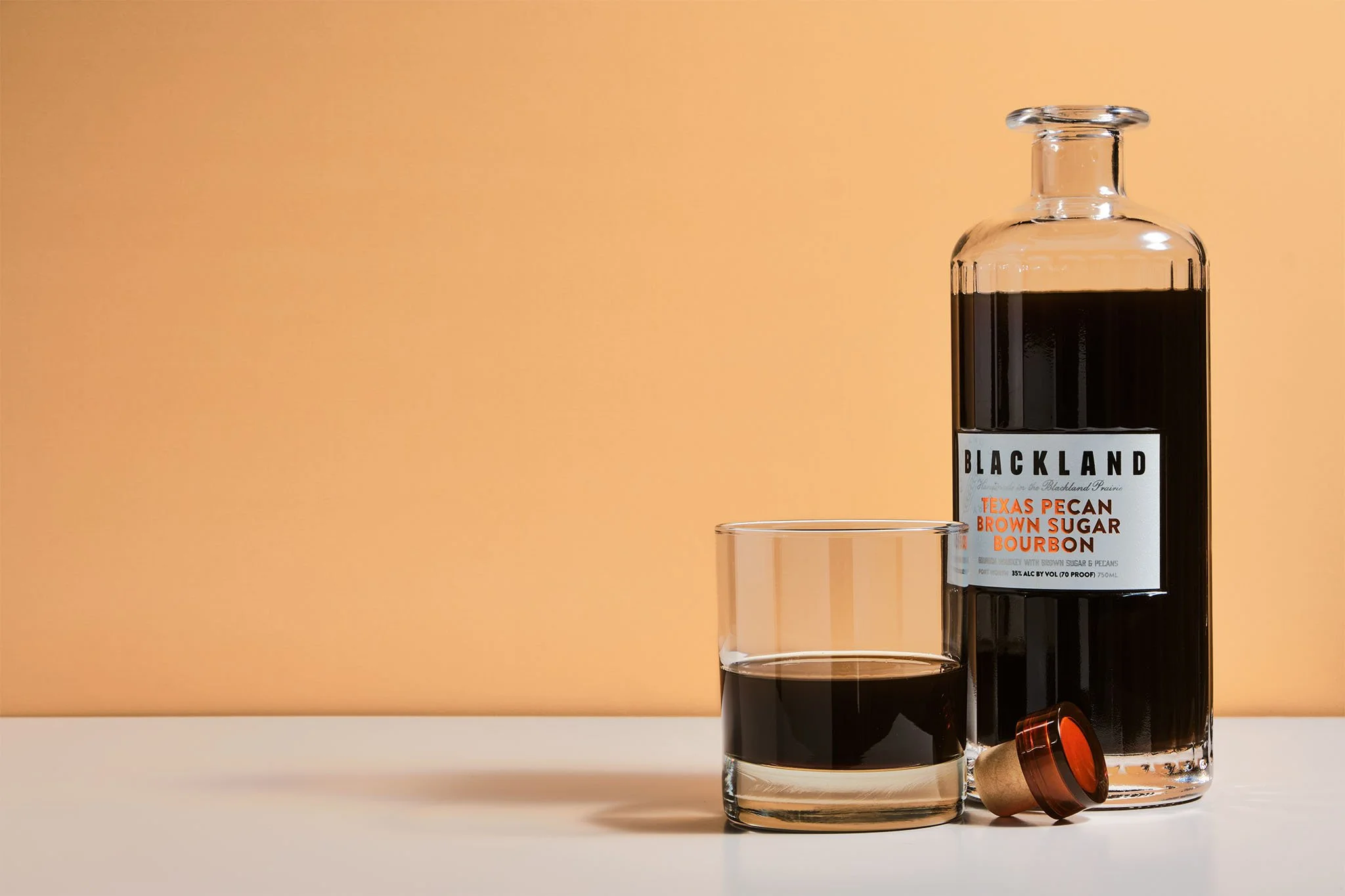 A glass bottle labeled 'Blackland Texas Pecan Brown Sugar Bourbon' with a glass of dark liquor in front of it, and a cork stopper lying next to the glass on a white surface against a peach-colored wall.