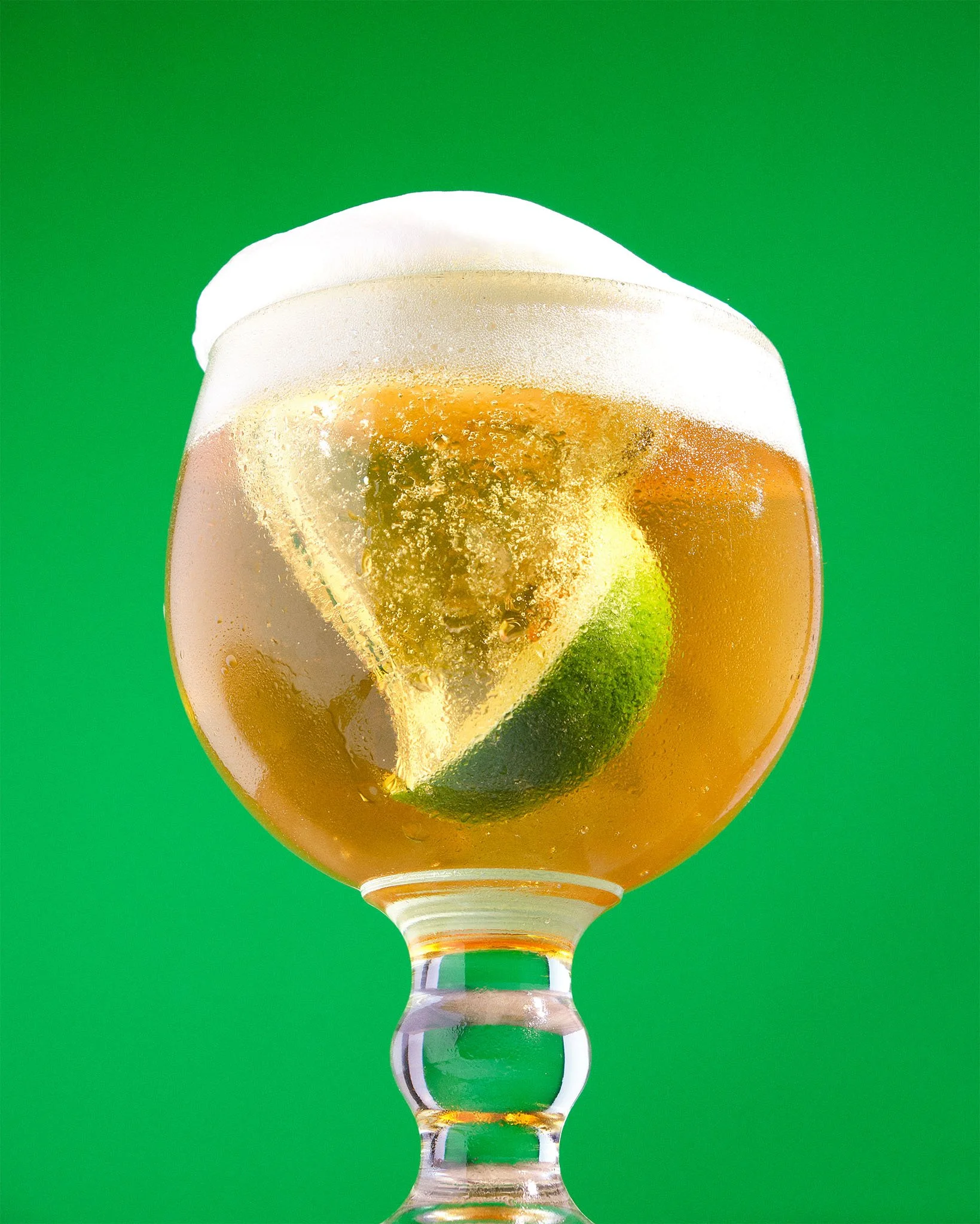 A glass of beer with a lime wedge and a frothy head against a green background.