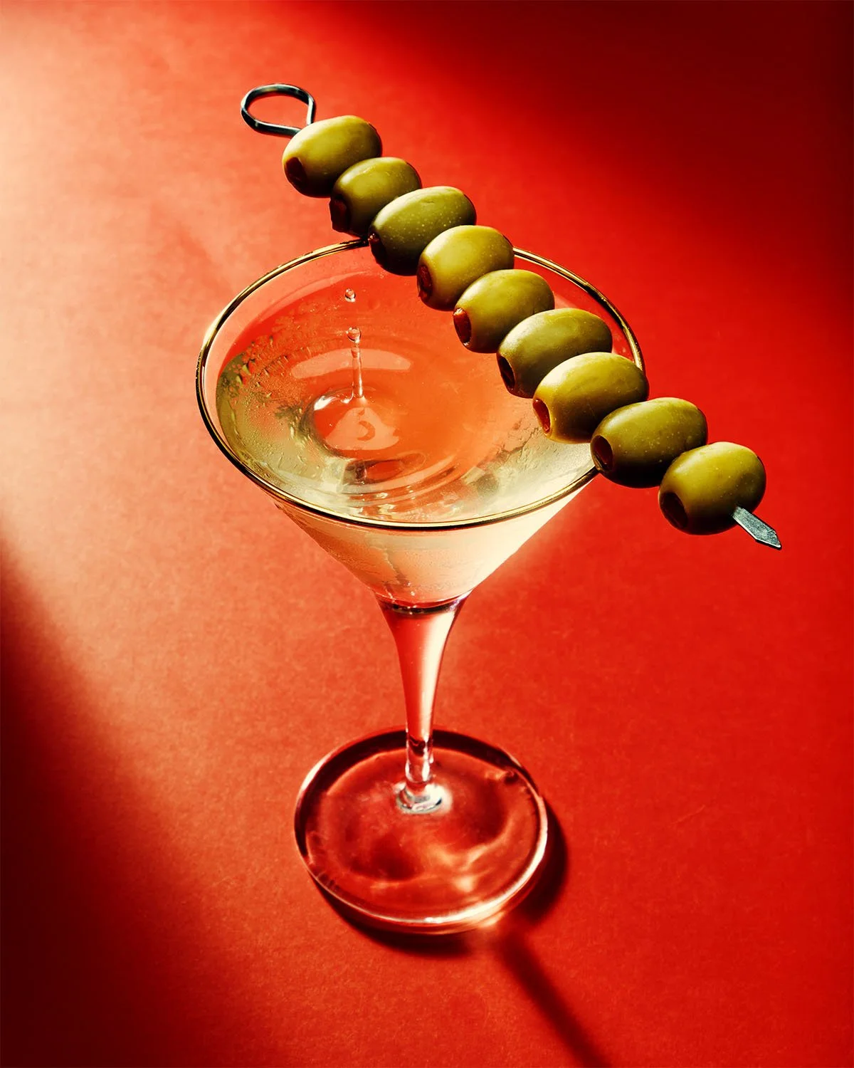 A martini glass with a light-colored cocktail, garnished with green olives threaded on a skewer resting on the glass rim, against a red gradient background.