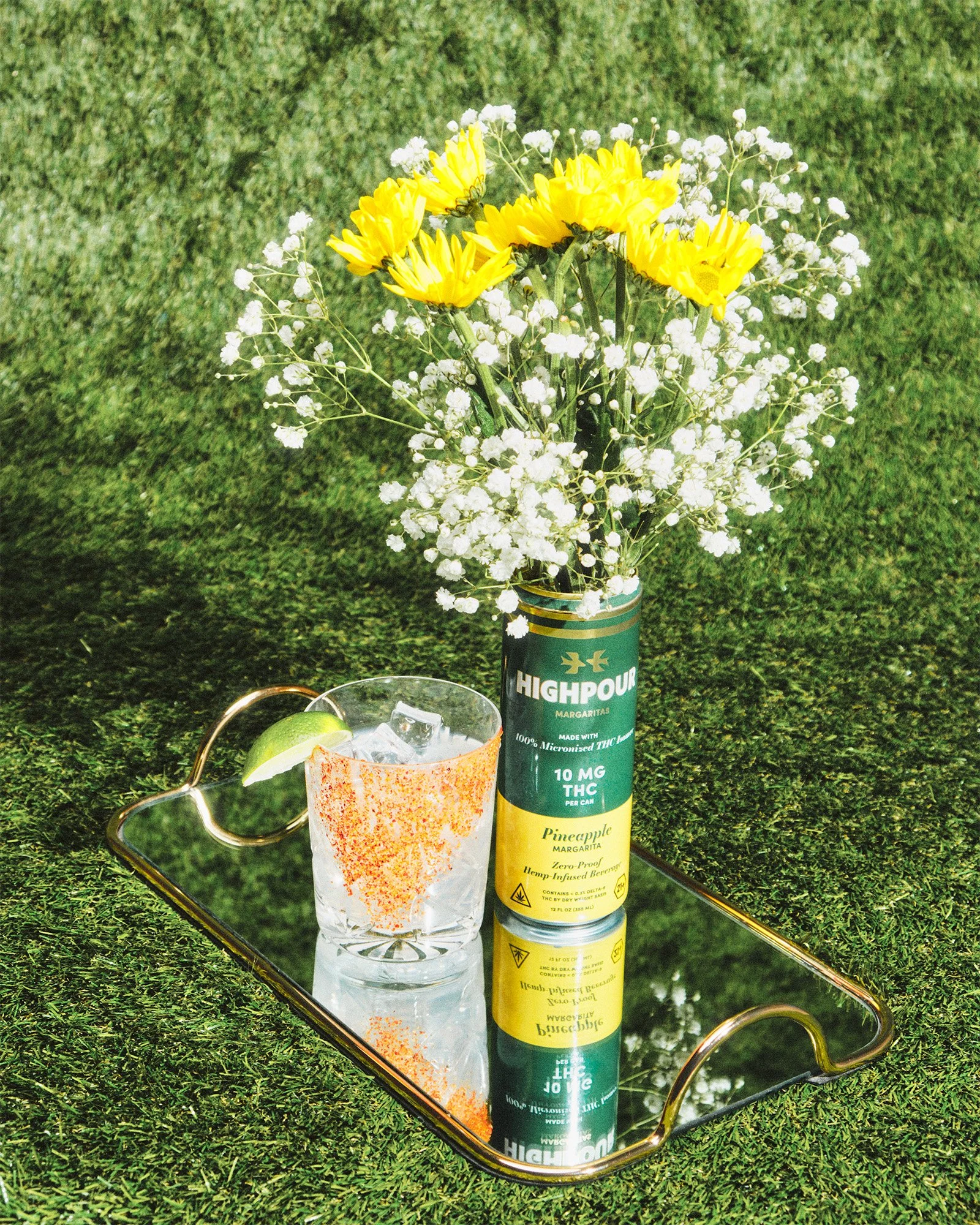 A bouquet of yellow and white flowers in a can labeled 'High Pour' resting on a tray. Nearby is a glass of ice with a lime wedge, set on a grassy surface.