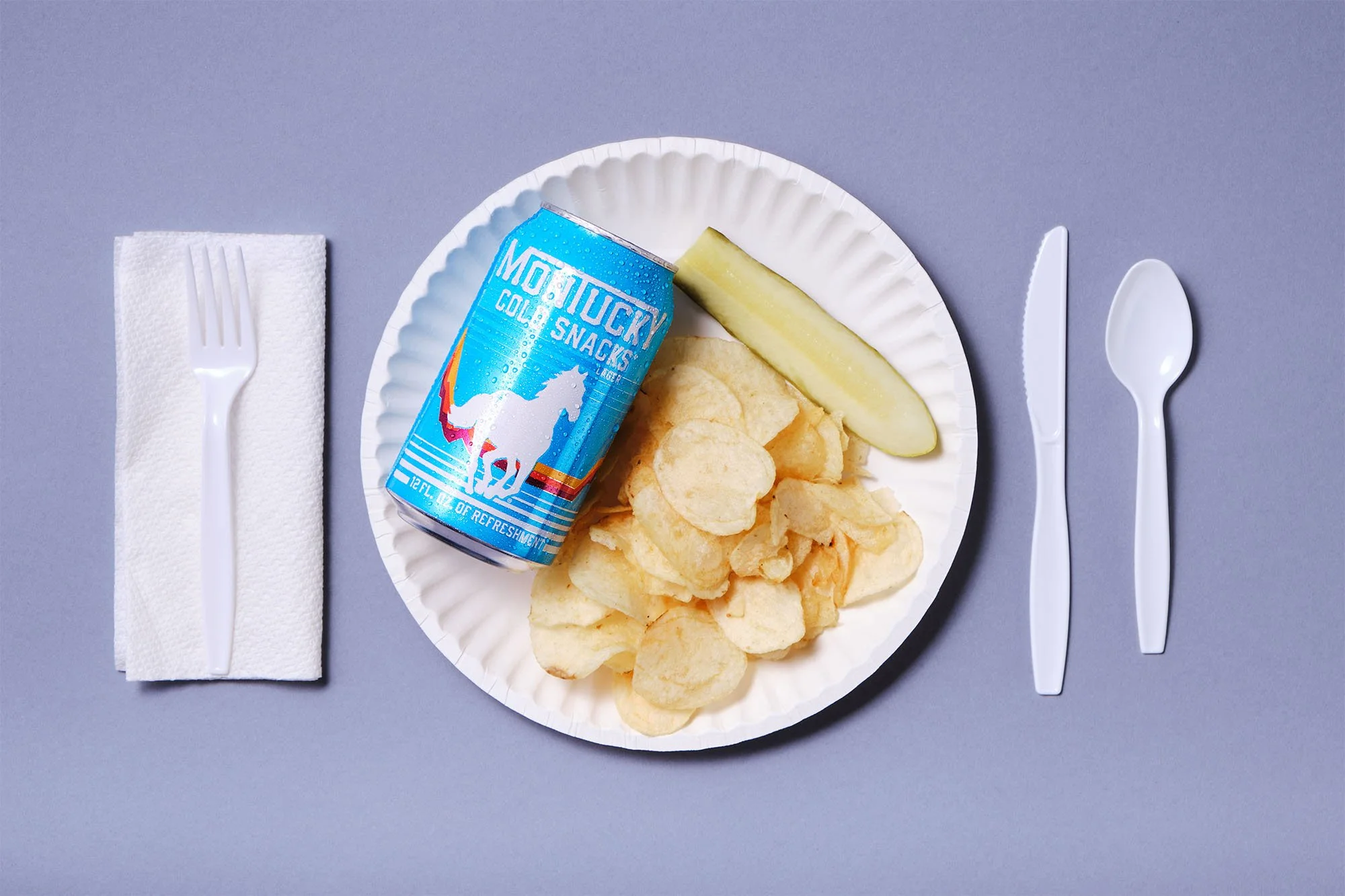 A paper plate contains potato chips, a slice of melon, and a can of Motuacky cold snacks. To the left of the plate is a white plastic fork on a paper napkin, and to the right are a plastic knife and spoon. The background is a solid light blue surface