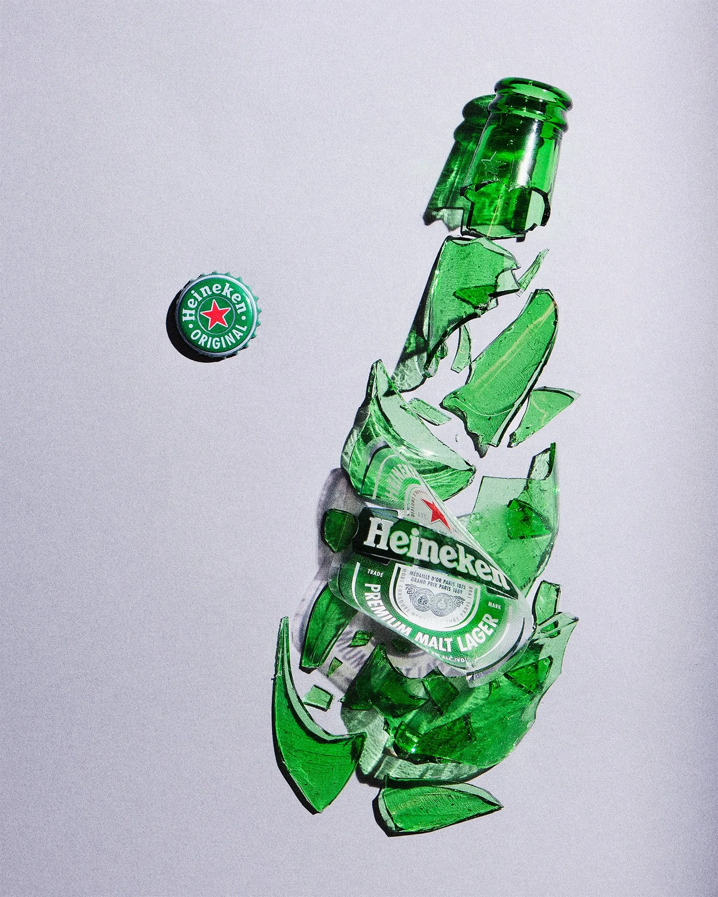 Broken green Heineken beer bottle with cap and label visible, fragments scattered on a white surface.
