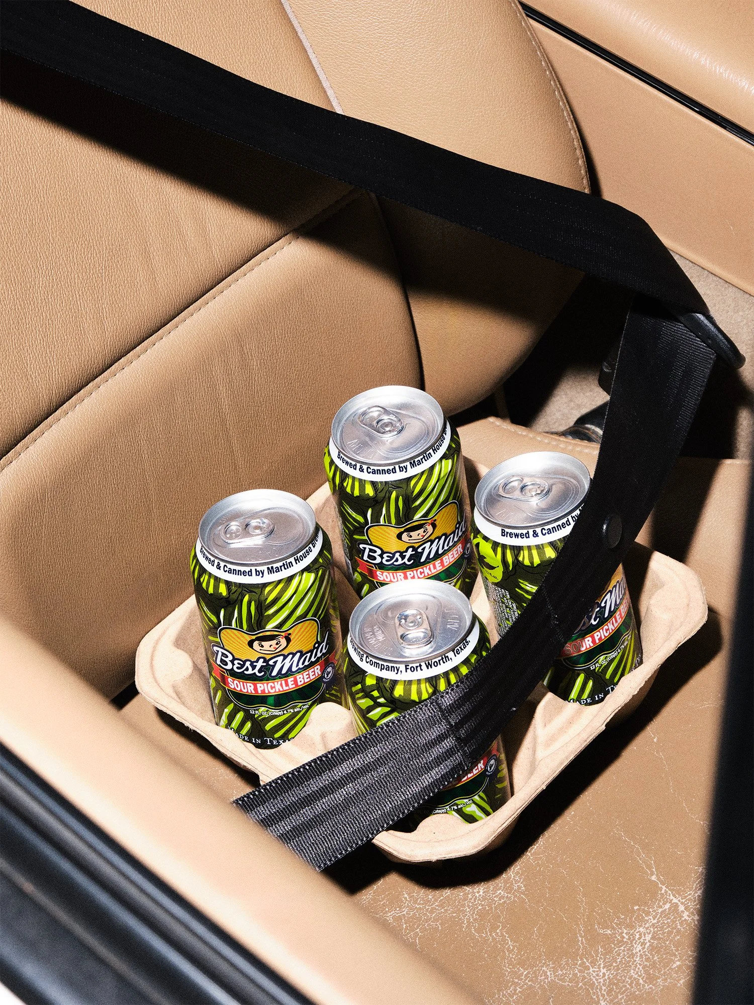 A carton containing six cans of Best Maid Sour Pickle Beer inside a car on a beige leather seat.