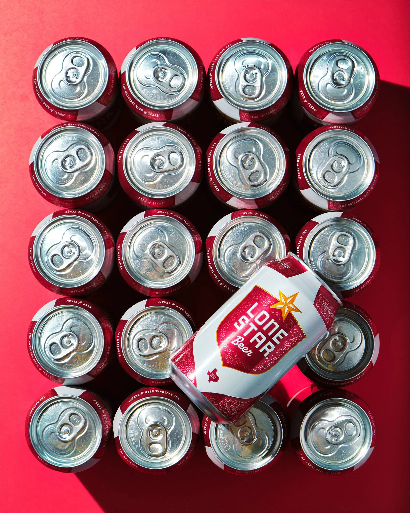 Top-down view of 16 cans of Lone Star Beer arranged on a red surface, with one can slightly tilted and prominently displayed.