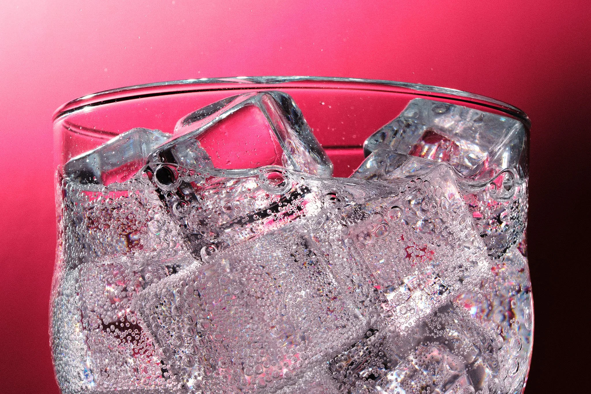 A glass filled with clear soda and ice cubes, with bubbles rising, against a pink background.
