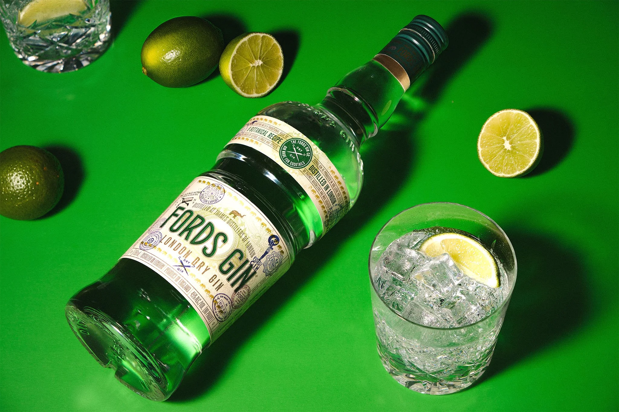 A bottle of Fords Gin lying on a green surface with half a lemon, a lime, and a glass of gin with ice and lemon slice, along with whole and cut limes in the background.