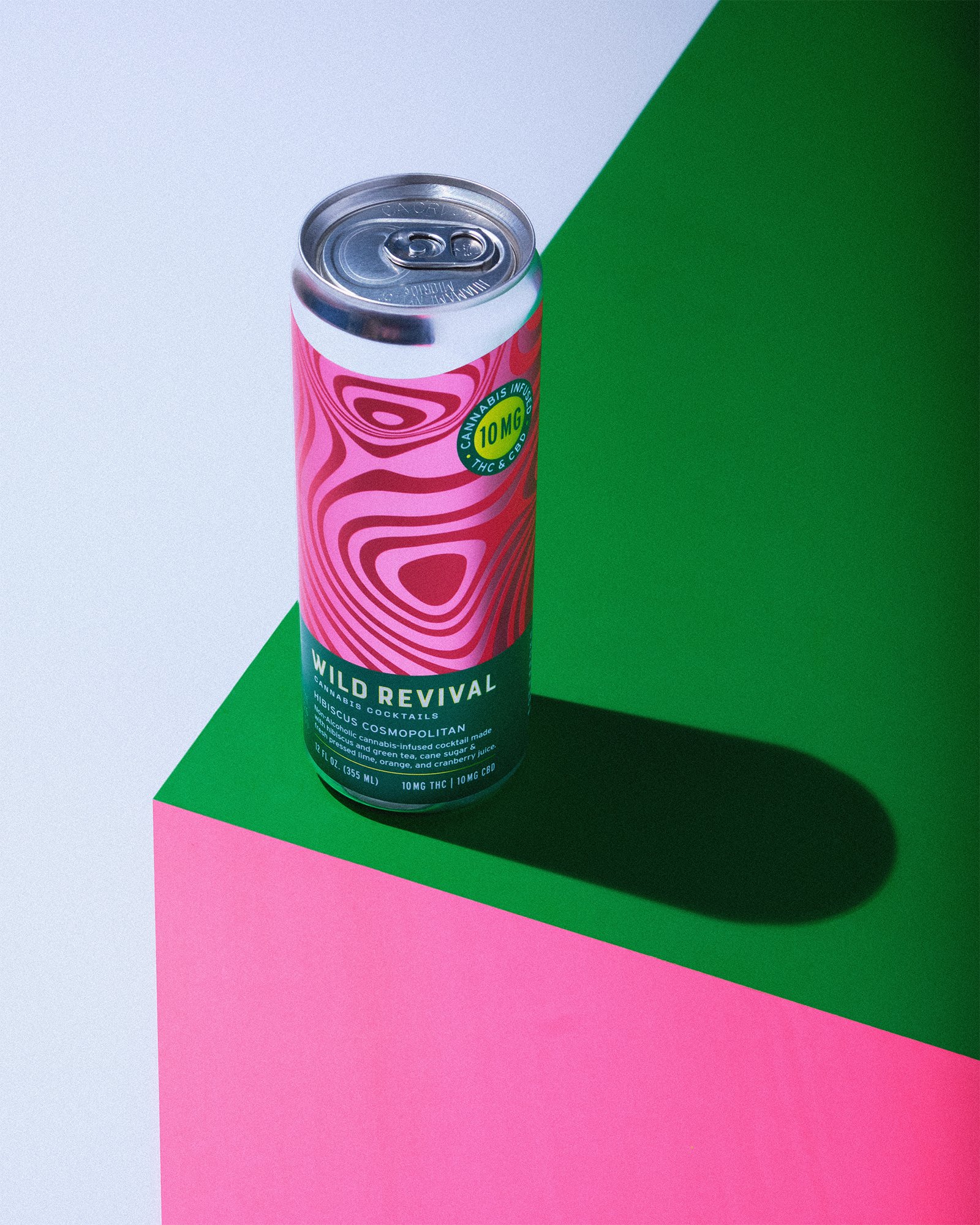 A pink and green can of cannabis-infused cocktail called Wild Revival with a wavy pink design on the label, placed on a multicolored geometric background of green, pink, and white.