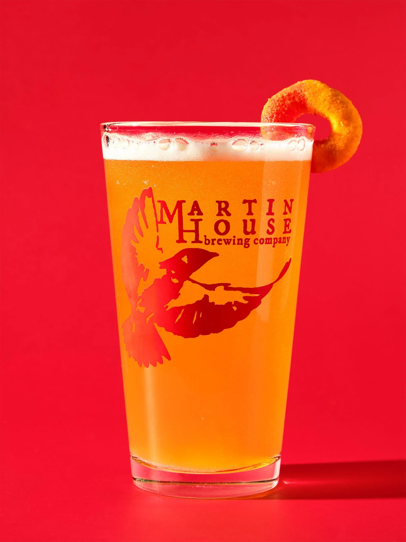 A glass of beer with an orange ring garnish, featuring the Martin House Brewing Company logo, against a red background.
