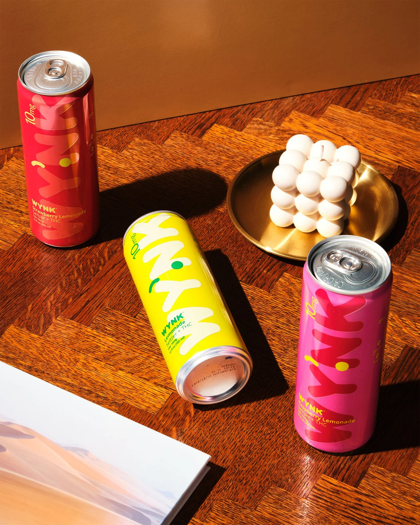 Two cans of WYNK Lemonade, one pink and one yellow, a golden dish with white ping pong balls, and part of a book on a wooden table.