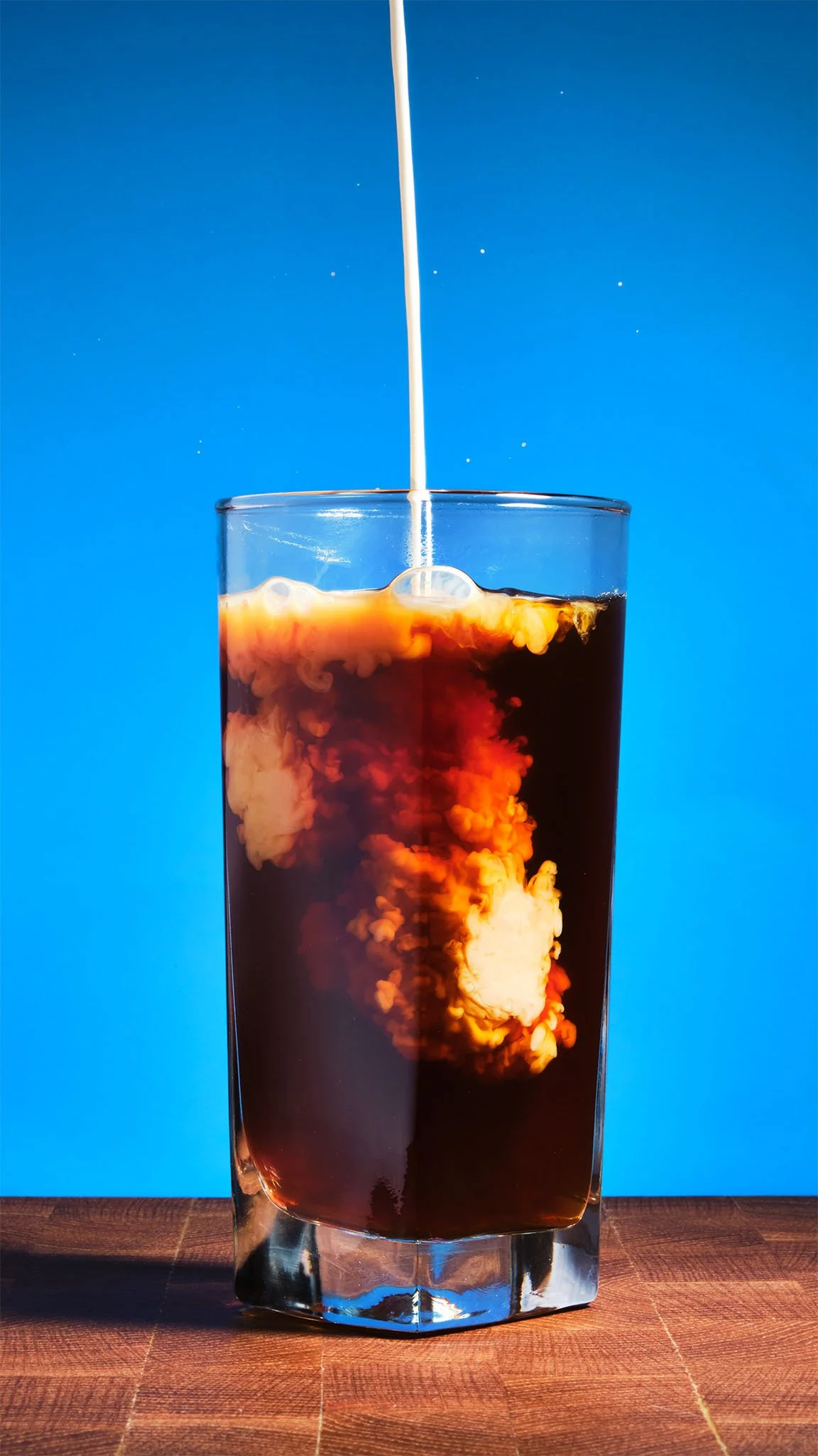 A tall glass of dark soda with cream being poured into it, creating a swirling effect, placed on a wooden surface with a blue background.