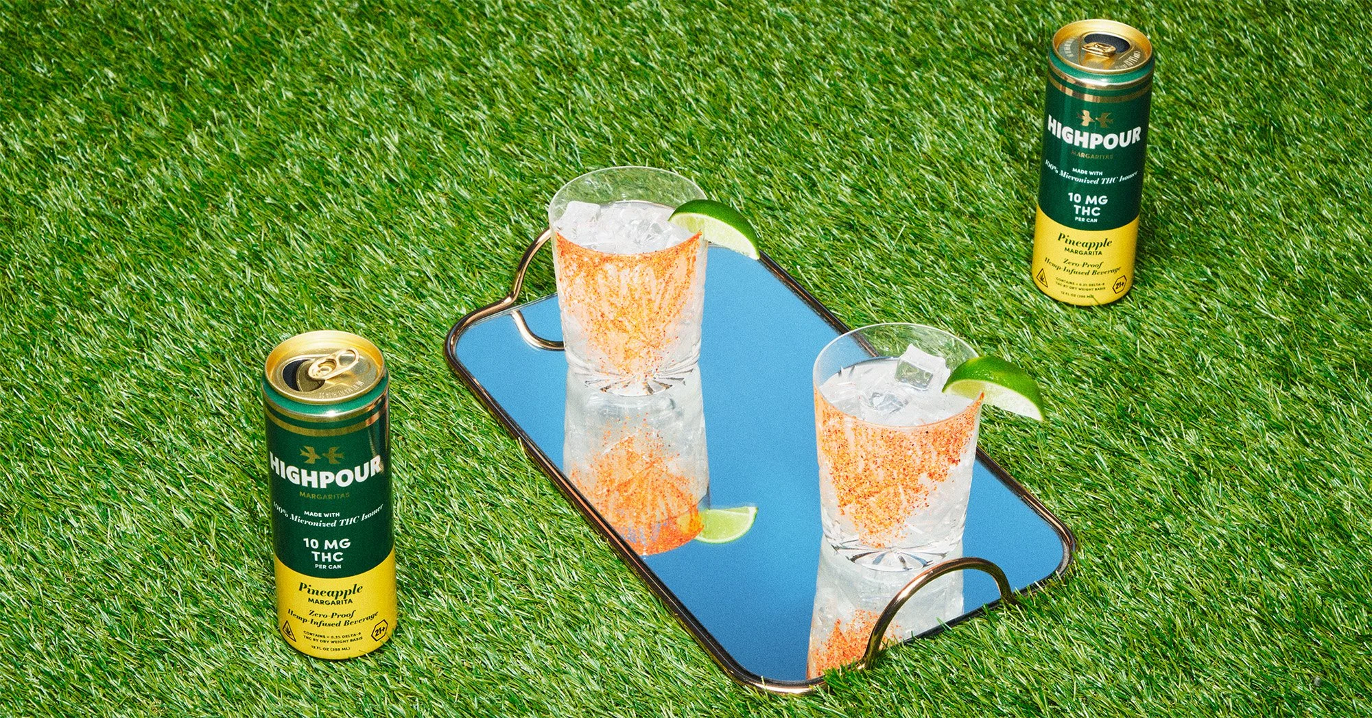 Two cans labeled 'Highpour' pineapple flavored THC-infused beverage are placed on artificial grass. A mirror on the grass reflects two tropical drinks garnished with lime wedges, ice, and salt-rimmed glasses, with one drink on a rectangular mirror tr