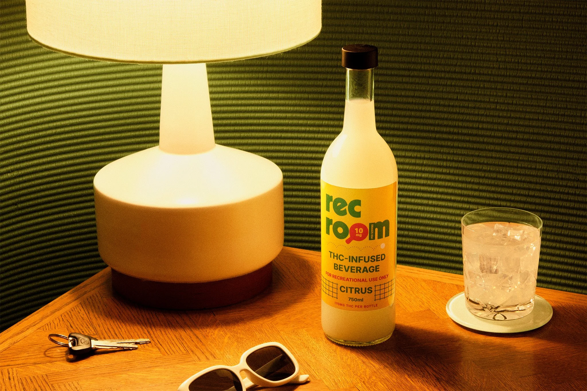 A table with a lamp, a bottle labeled THC-infused beverage, a glass of water with ice, a pair of sunglasses, and a set of keys. The background has a green textured wall.