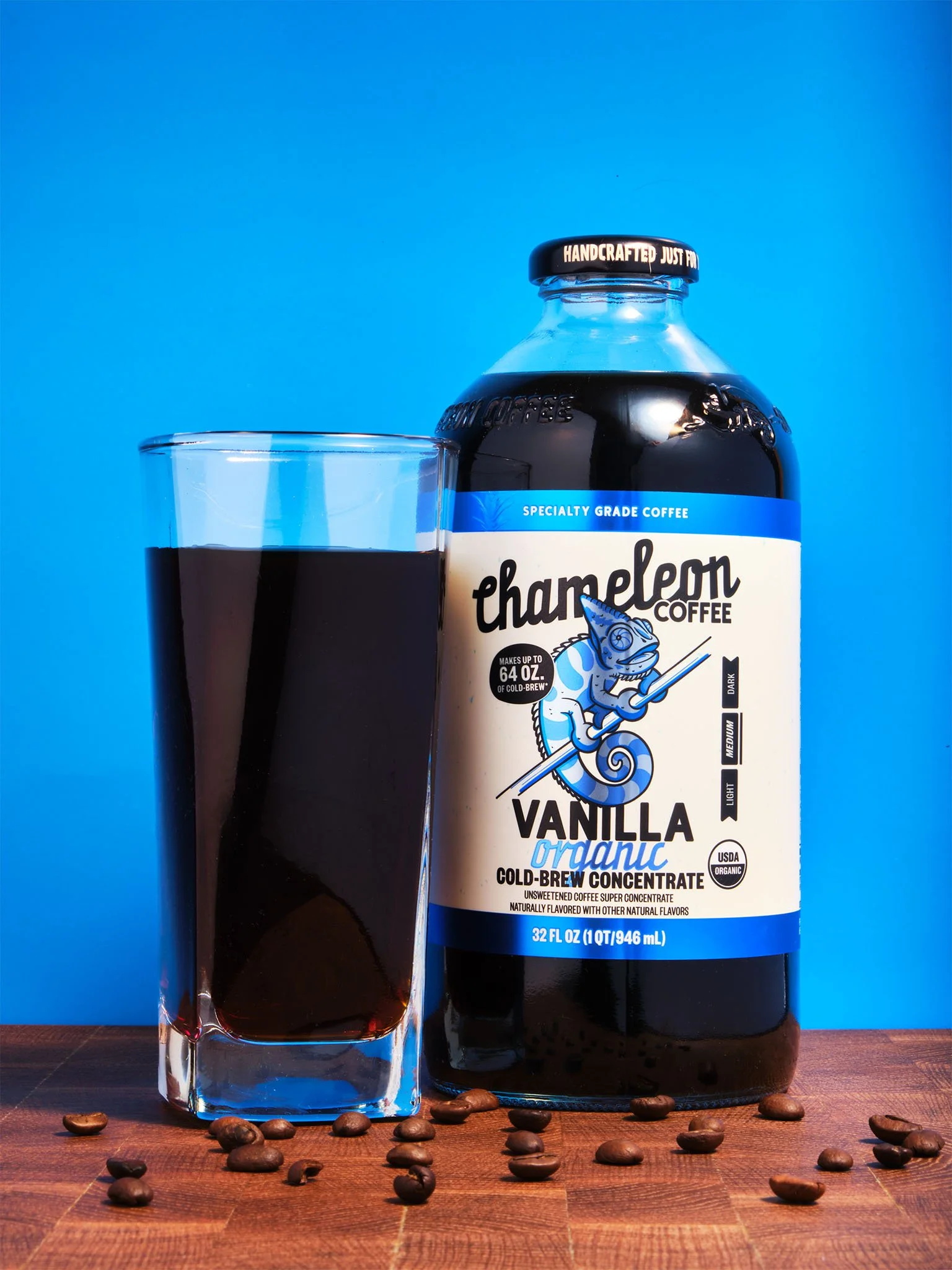 A glass of cold brew coffee next to a large bottle of Chamelon Coffee Vanilla organic cold-brew concentrate on a wooden surface, with scattered coffee beans in the foreground and a bright blue background.