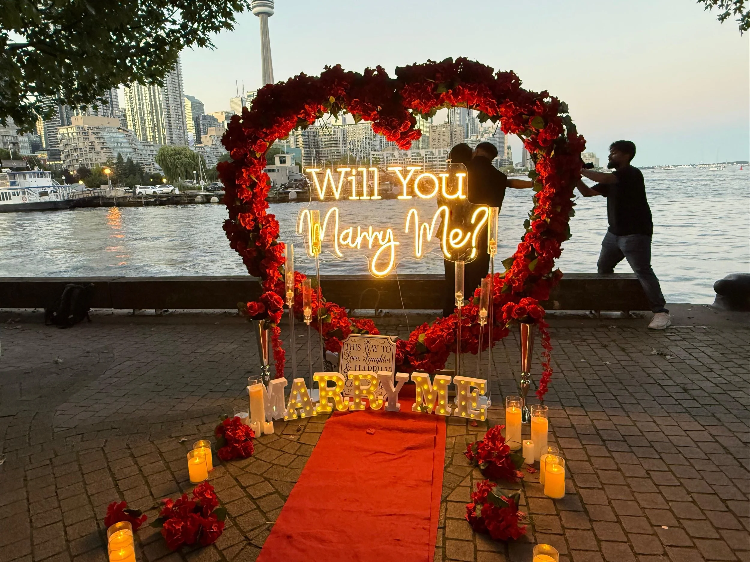 A romantic proposal setup by the water with a heart-shaped floral decoration, illuminated signs saying "Will You Marry Me?" and "Marry Me," surrounded by candles and a red carpet, with city buildings and a skyline in the background at sunset.