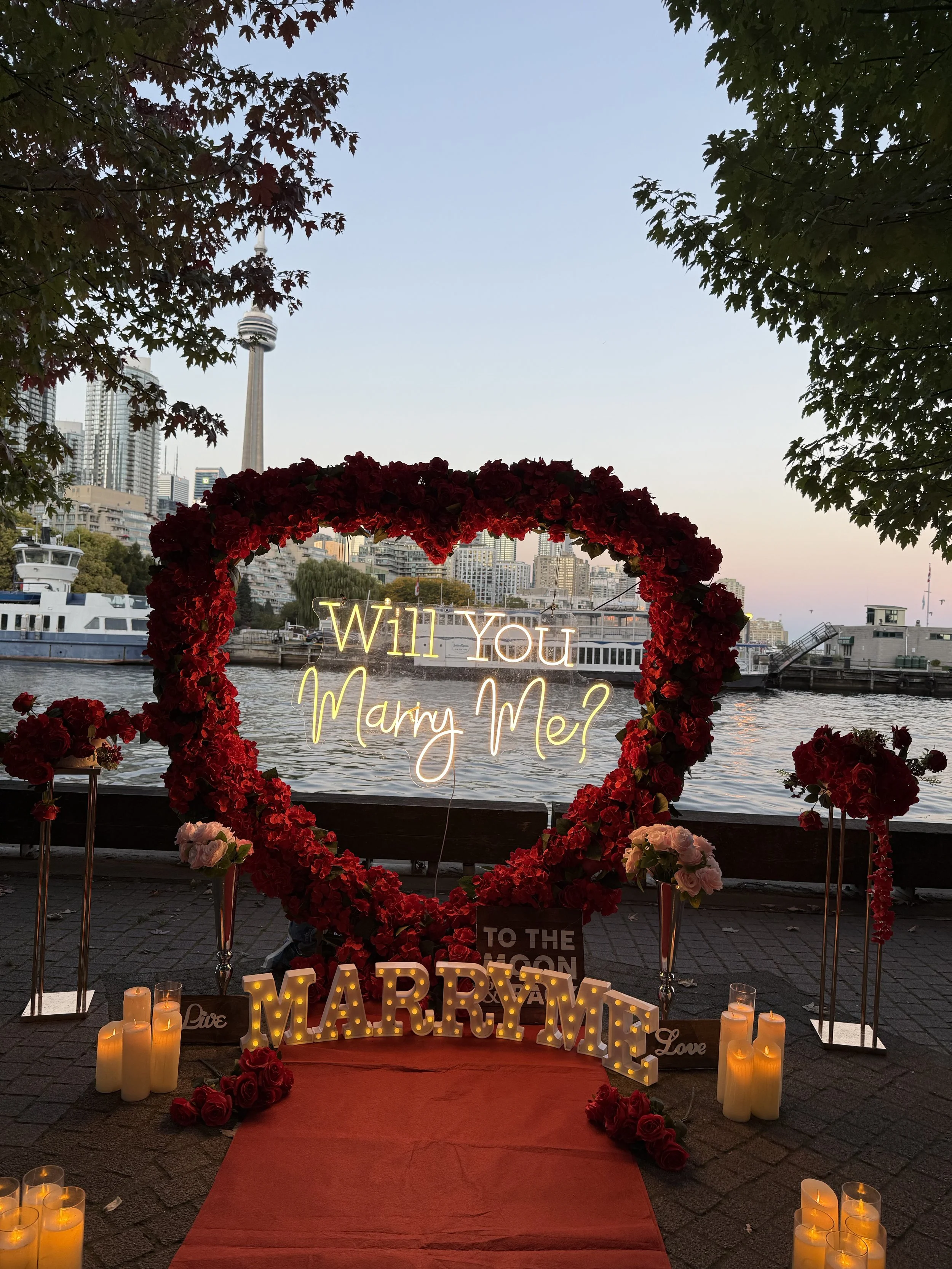 Outdoor romantic proposal setup by a river featuring a large heart-shaped floral arrangement, illuminated neon sign asking "Will You Marry Me?", lit marquee spelling out "MARRY ME", candles, roses, and city skyline in the background at dusk.