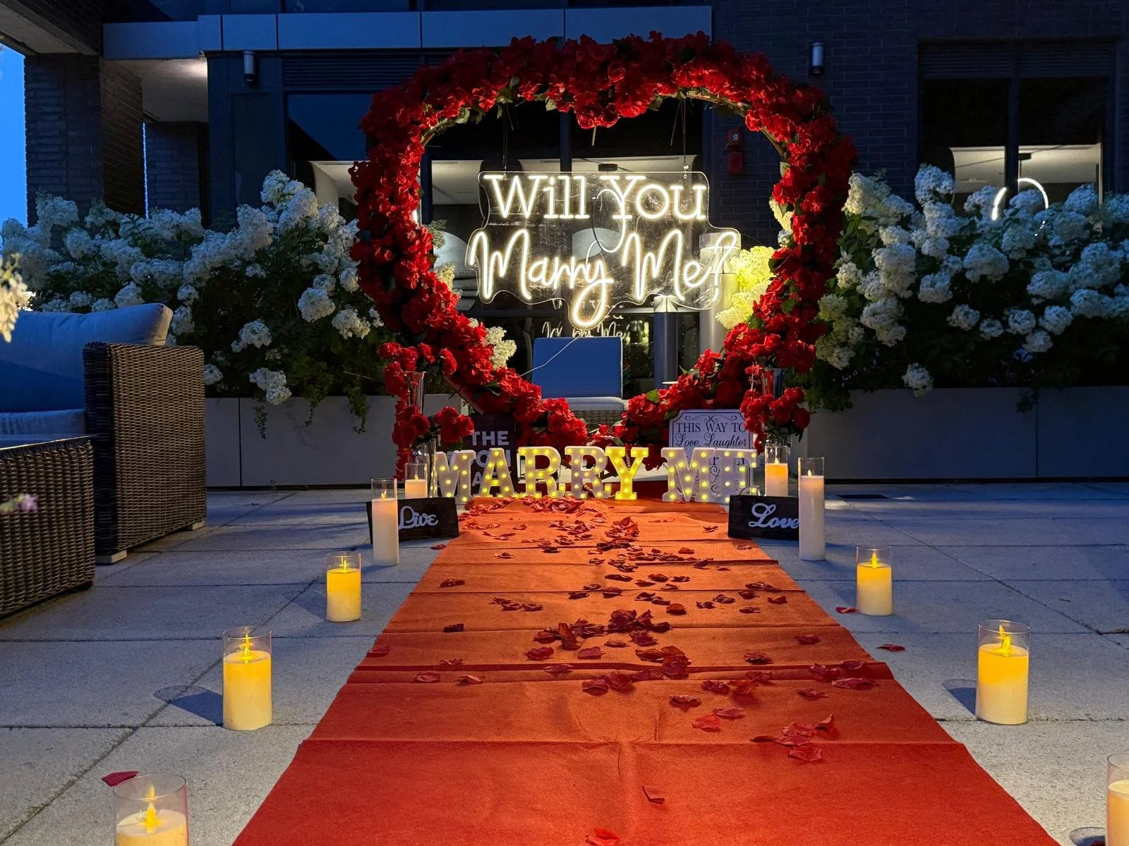 Outdoor wedding proposal setup with a red carpet, candles, and flower decorations, including a heart-shaped flower arrangement with a neon sign that says 'Will You Marry Me?'