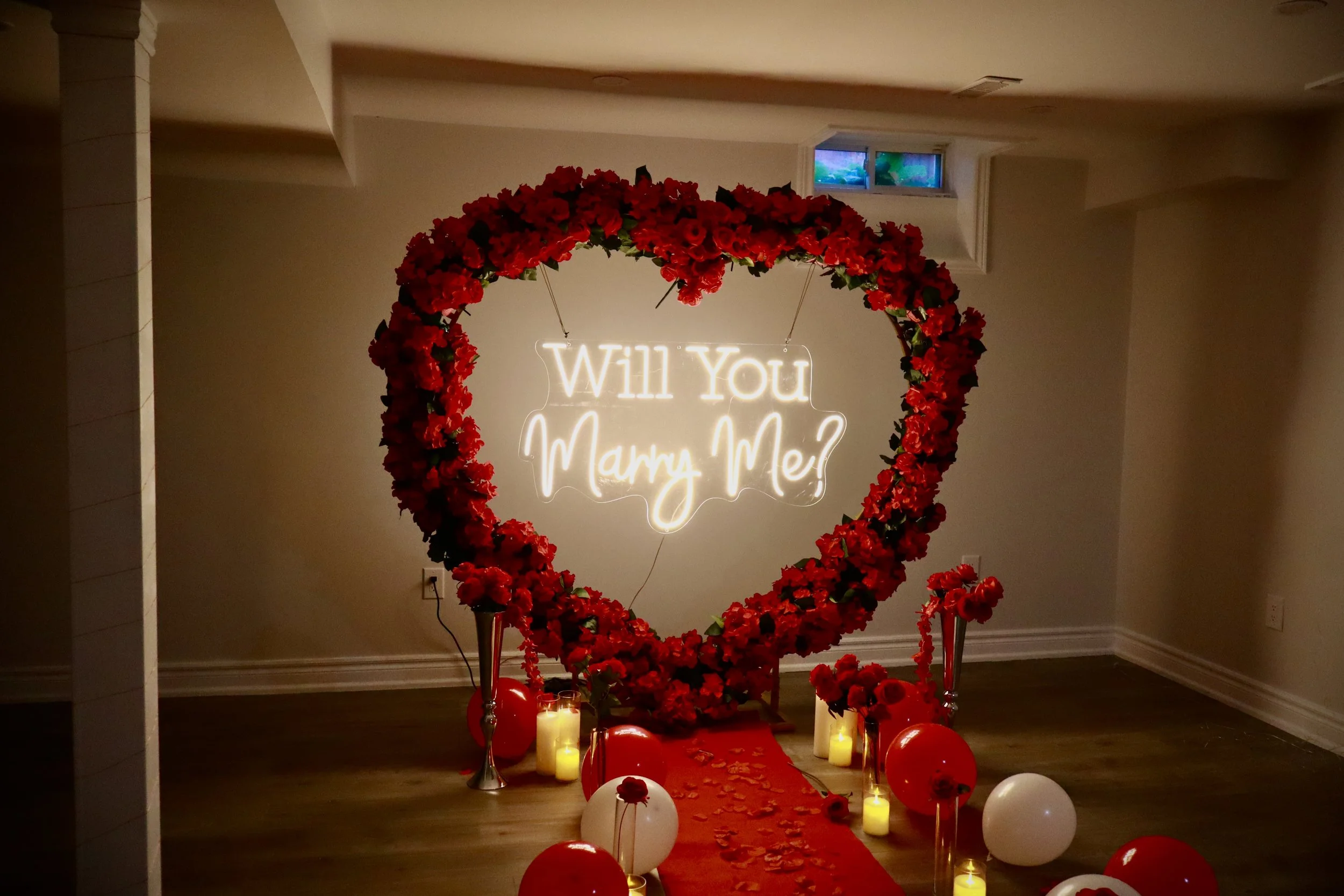 A romantic proposal setup with a heart-shaped flower arrangement, a neon sign asking 'Will You Marry Me?', surrounded by candles and balloons on the floor.
