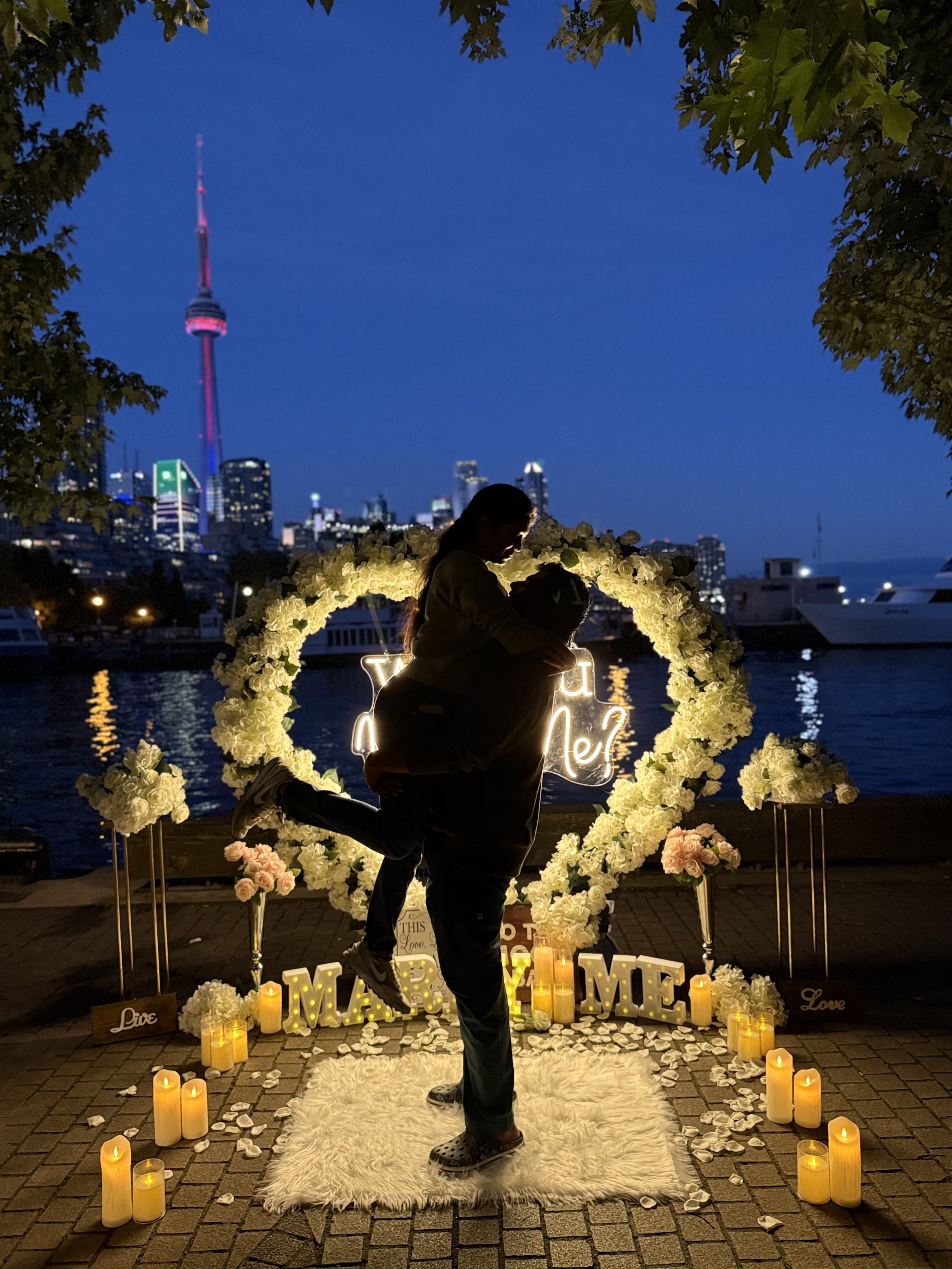 A silhouetted couple is kissing on a white furry rug surrounded by candles and flower arrangements, with a city skyline and a body of water in the background, during evening or nighttime.