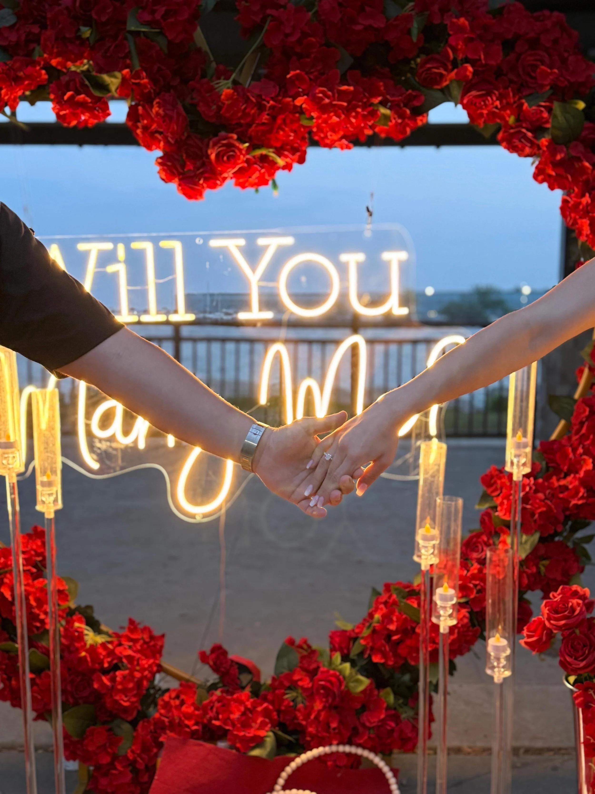 A couple holding hands with wedding rings, surrounded by red flowers and a neon sign that says 'I will you' in the background.