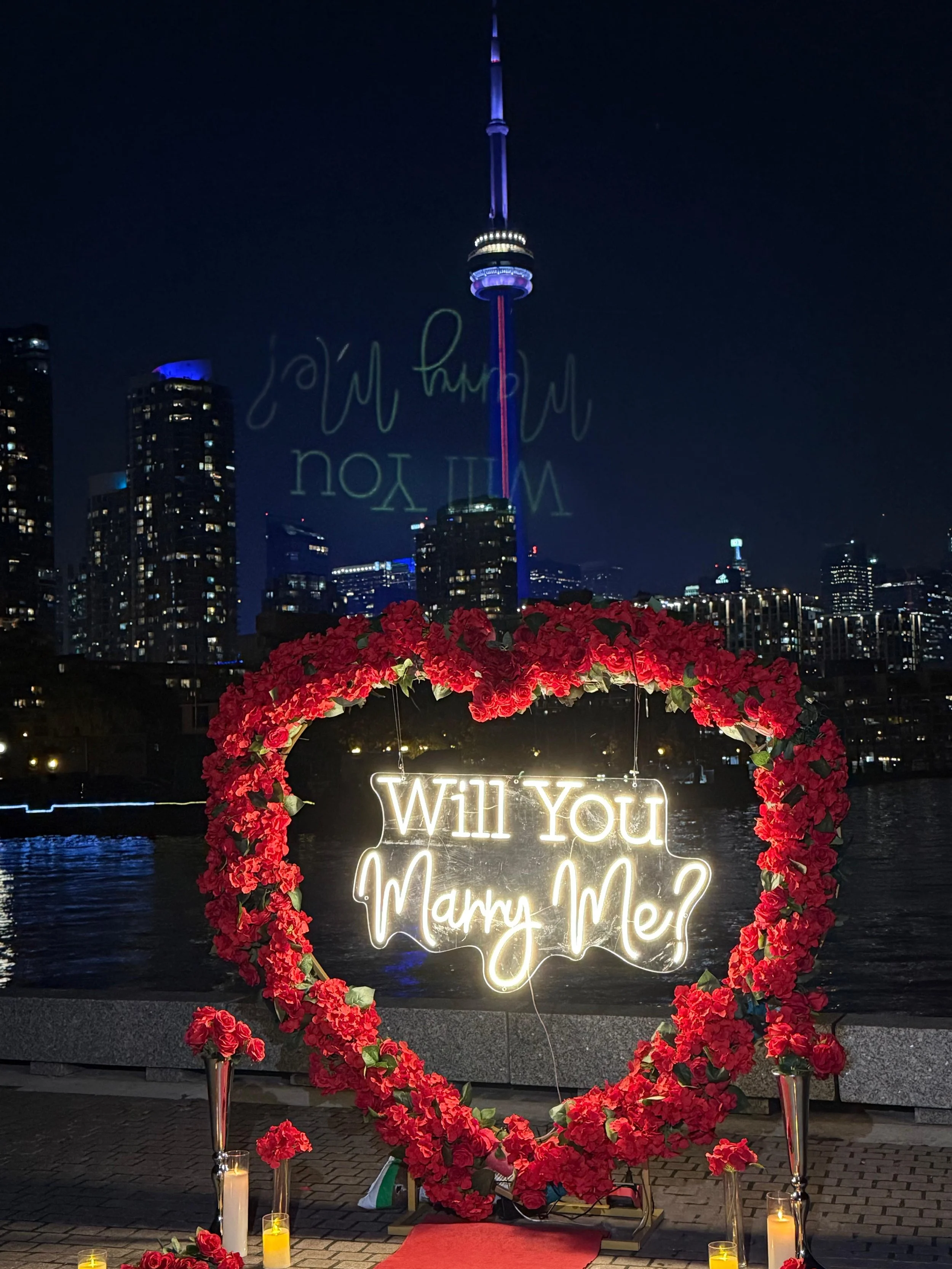 A neon sign that reads 'Will You Marry Me?' in front of a city skyline at night, surrounded by red flowers, candles, and a red carpet.