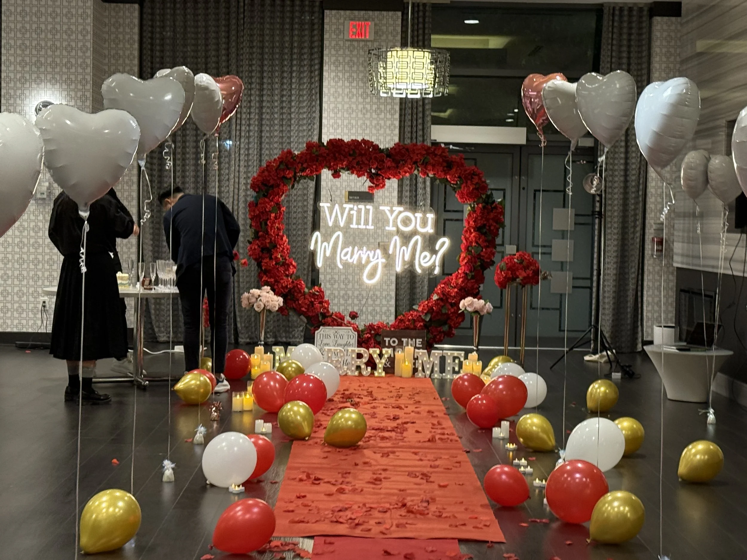 A romantic proposal setup with a heart-shaped floral arch, illuminated sign asking 'Will You Marry Me?', red carpet, red, white, and gold balloons, candles, and flower arrangements at an indoor venue.