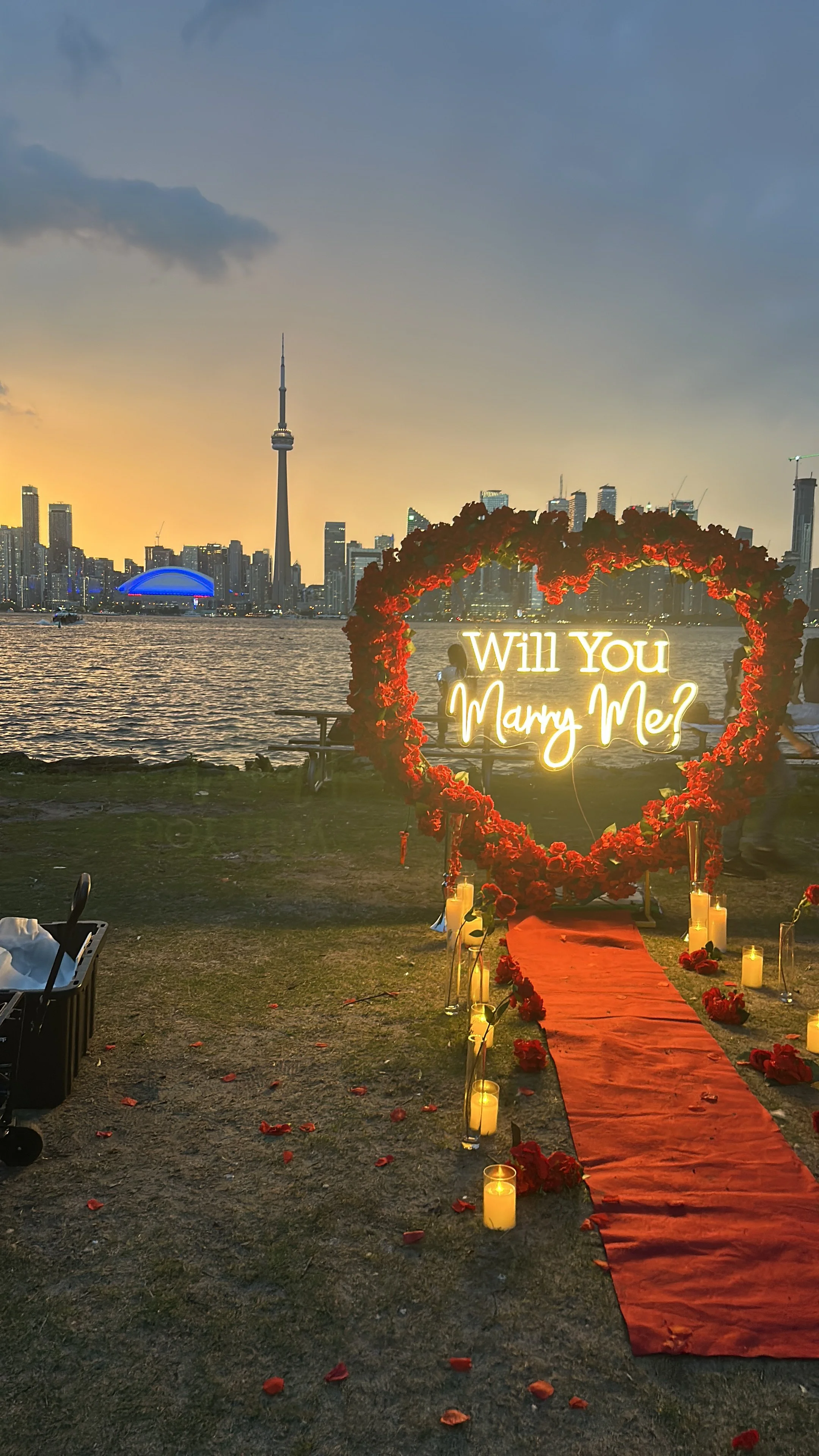 A romantic proposal setup on a waterfront with a city skyline, including the CN Tower, during sunset. The scene features a red carpet, candles, roses, and a neon sign inside a heart-shaped floral frame that reads, 'Will You Marry Me?'